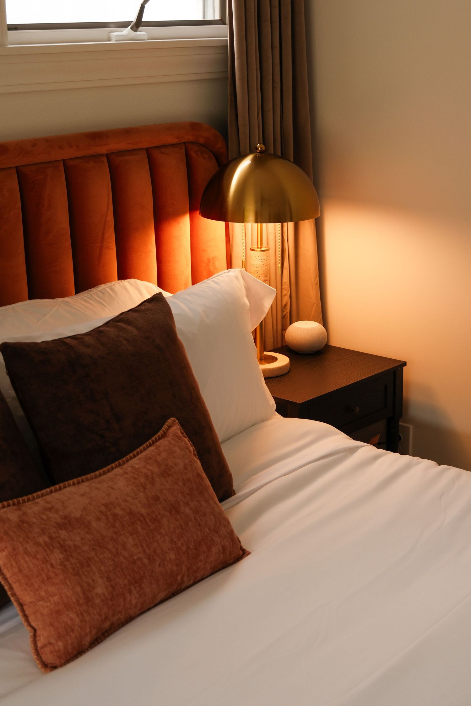 Cozy bedroom with rust velvet headboard, brown and white pillows, white bedding, and glowing brass lamp on dark nightstand.
