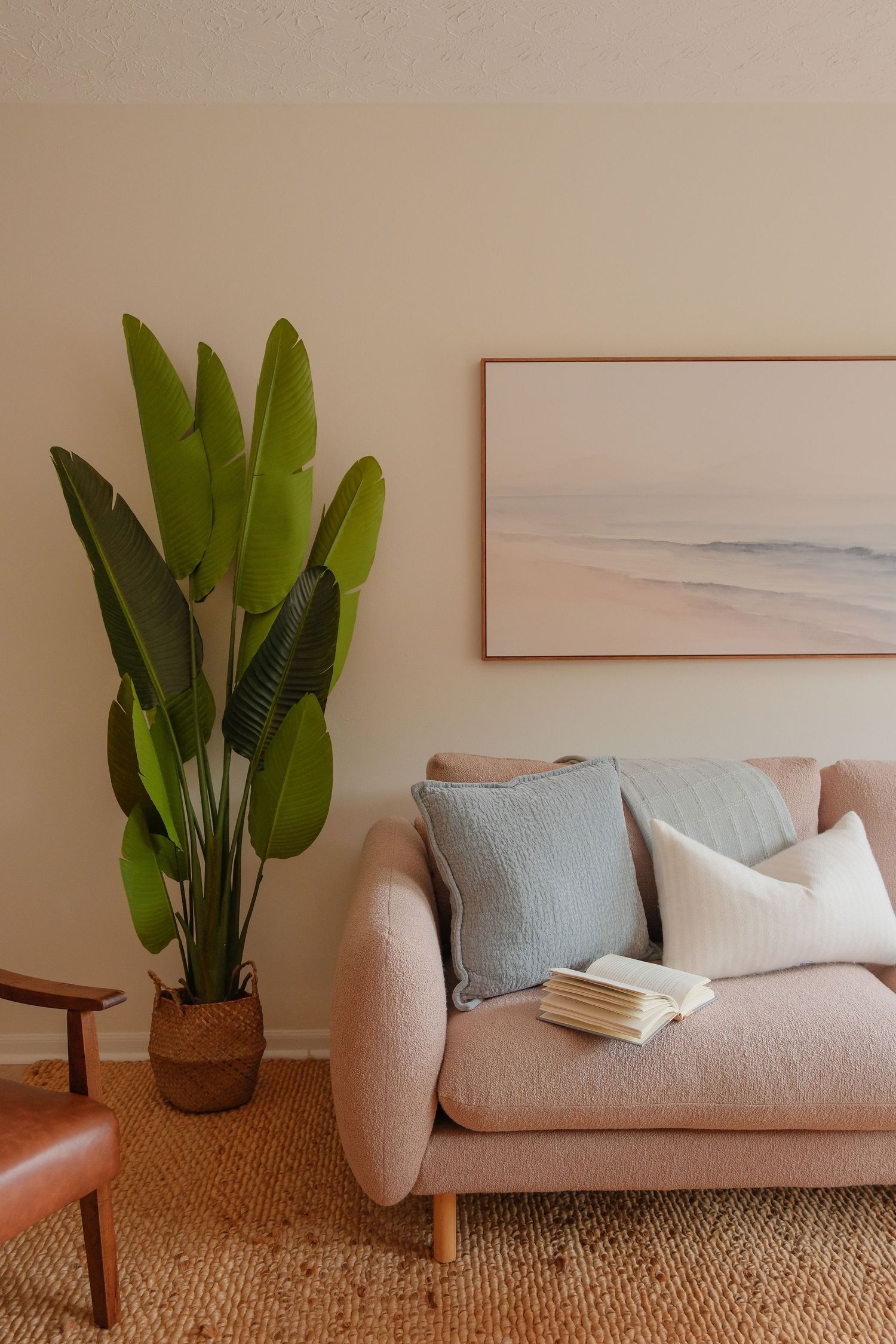 Cozy living room with pink sofa, blue pillows, open book, tall tropical plant, and serene beach artwork on beige wall.