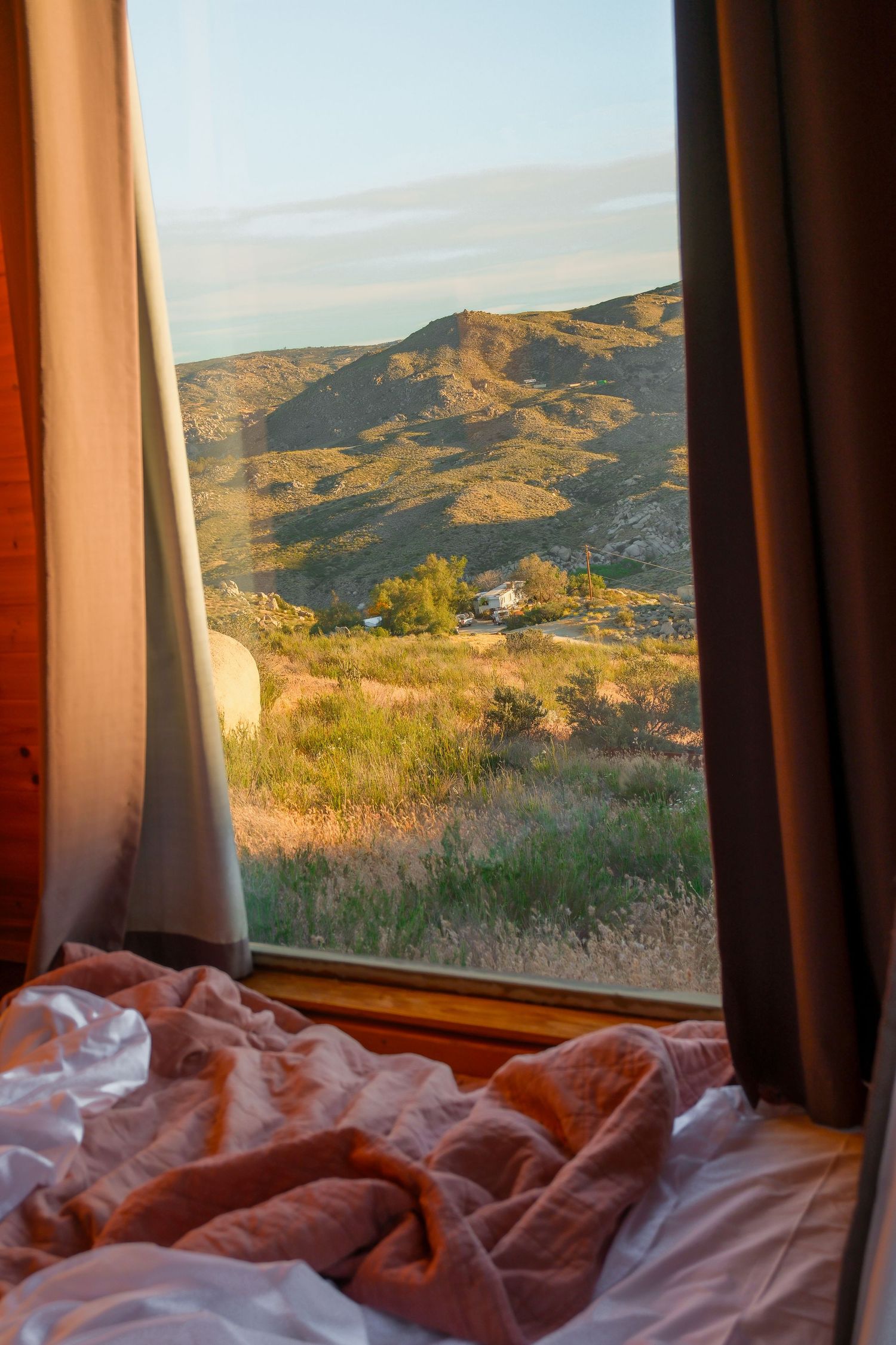 Cozy unmade bed with pink sheets beside an open window overlooking a scenic mountain valley at golden hour.