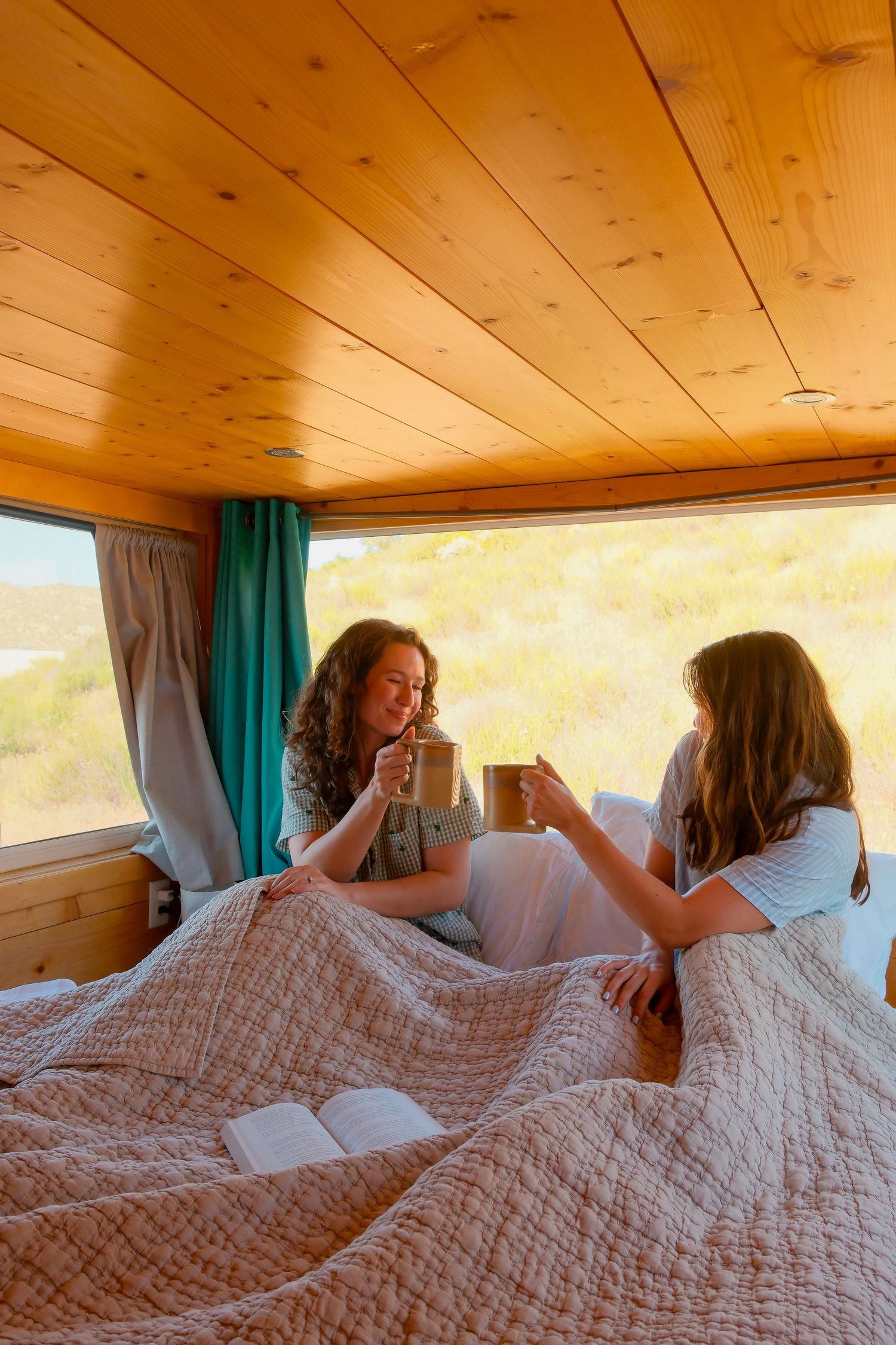Two women sitting in bed inside a wood-paneled camper van, drinking coffee with teal curtains and nature views.