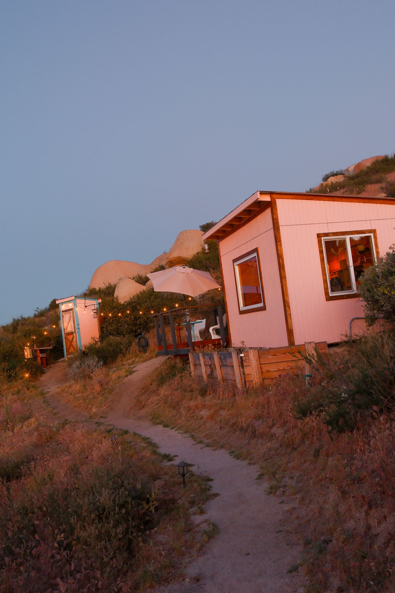 Small pink cabin with string lights along a winding dirt path on a hillside at dusk in a desert retreat.