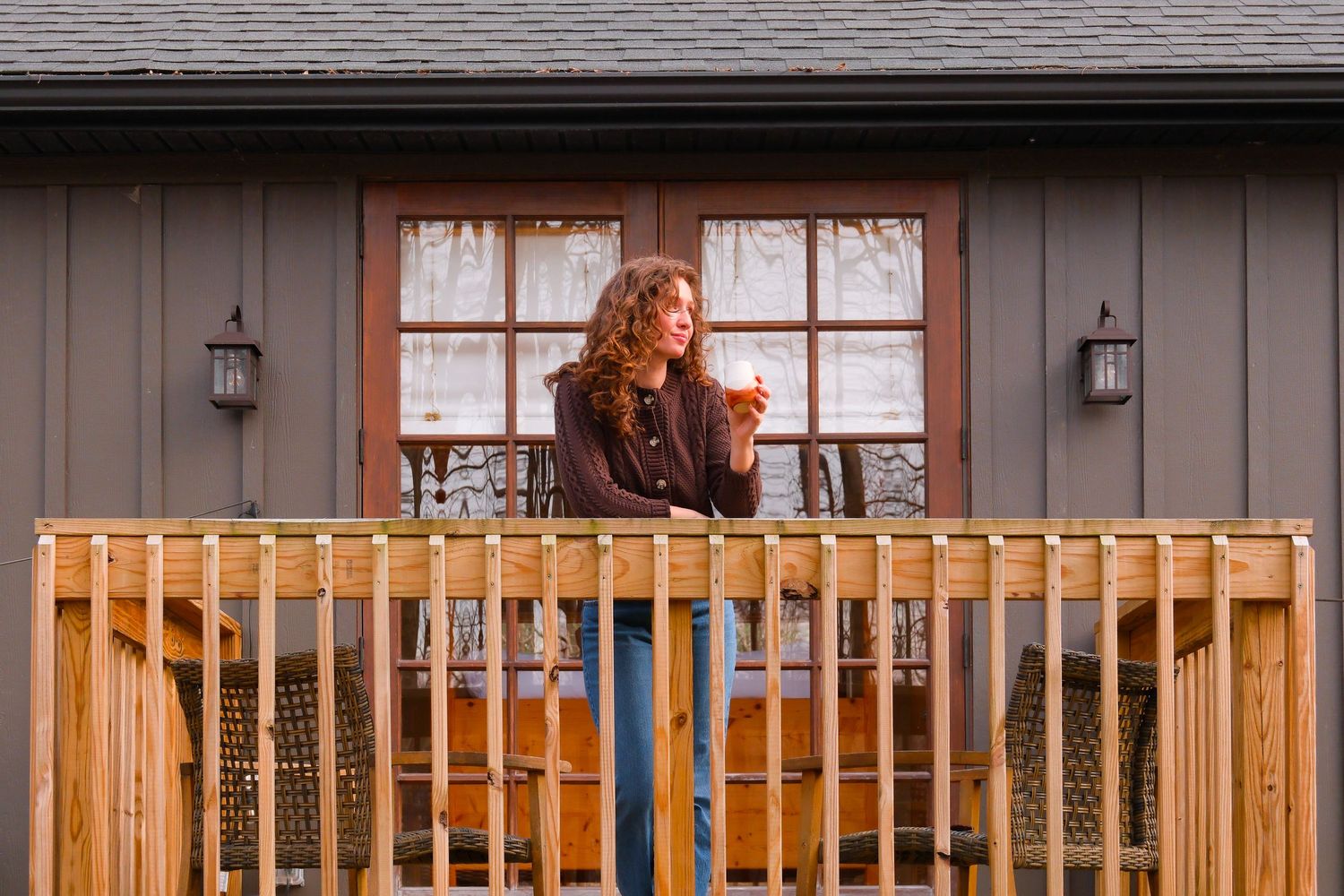 Woman relaxing on a wooden deck with a warm drink outside a charming gray cabin home.
