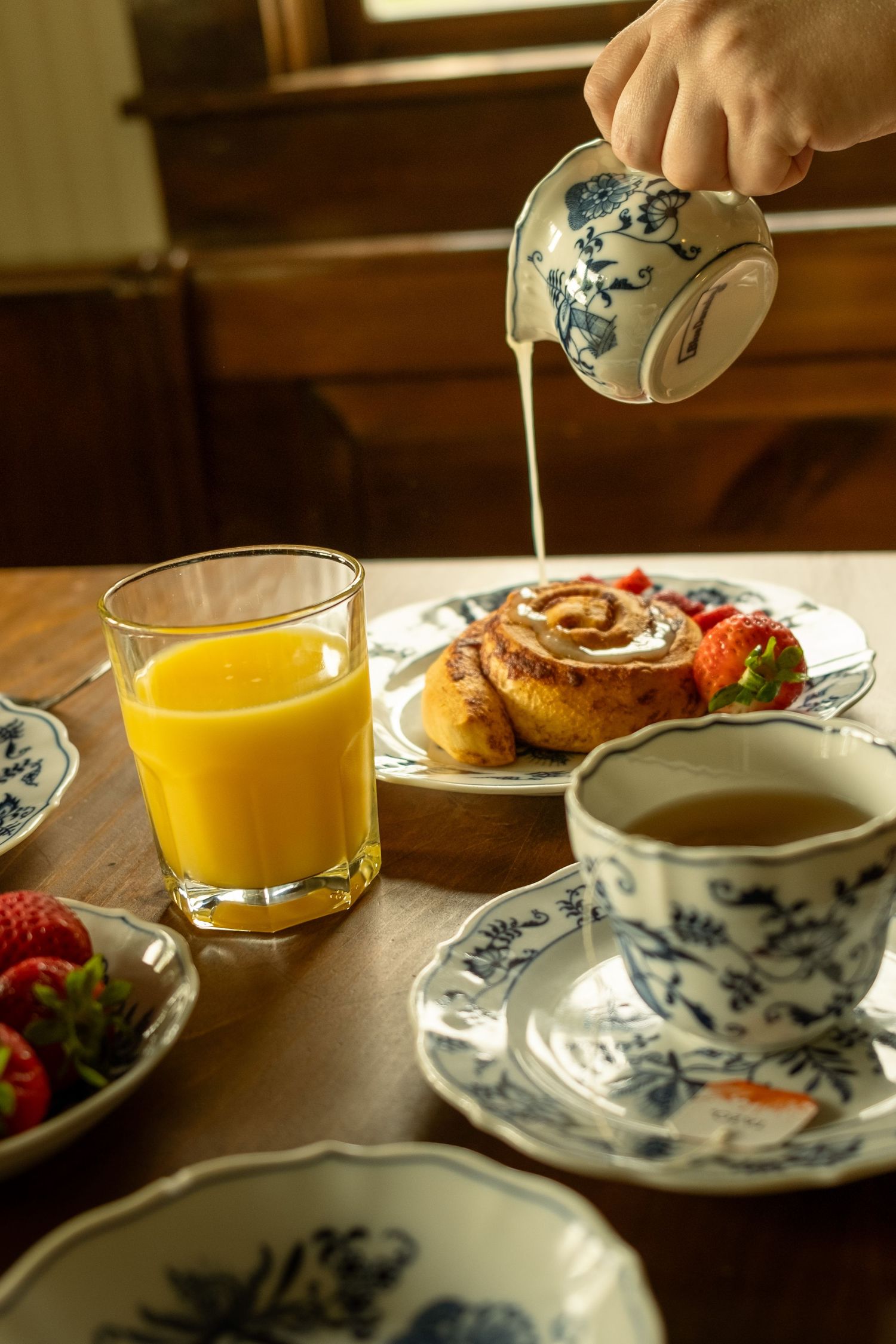 Pouring cream into coffee with breakfast plates of pastries and fruit in morning light.