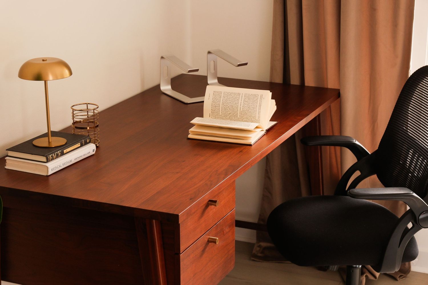 A wooden desk with an open book, gold lamp, and black office chair in a cozy study room.