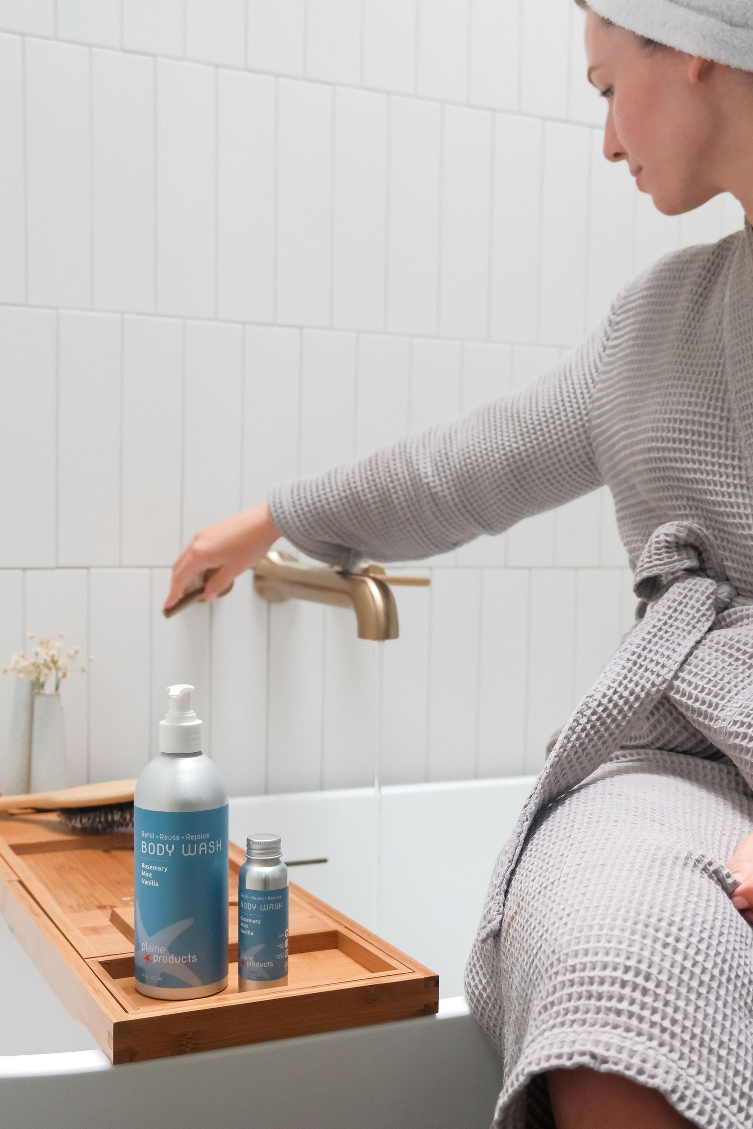 Woman in gray waffle robe sitting by bathtub with body wash products on bamboo tray.