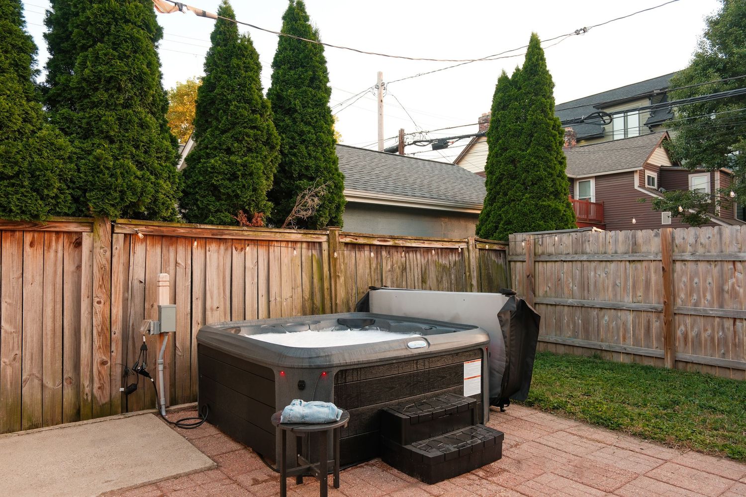 Backyard patio with a large hot tub, BBQ grill, surrounded by tall cedar trees and wooden privacy fencing.