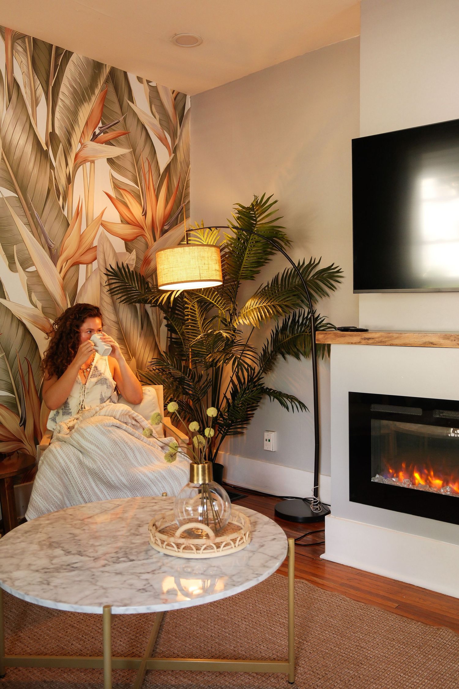 Woman relaxing in cozy tropical-themed living room with fireplace, palm wallpaper, and marble coffee table.