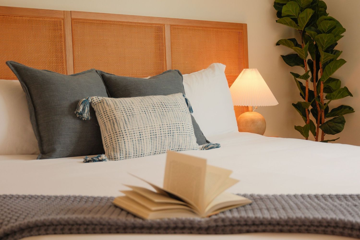 Cozy bedroom with white bedding, gray pillows, wooden headboard, and an open book resting on the bed.