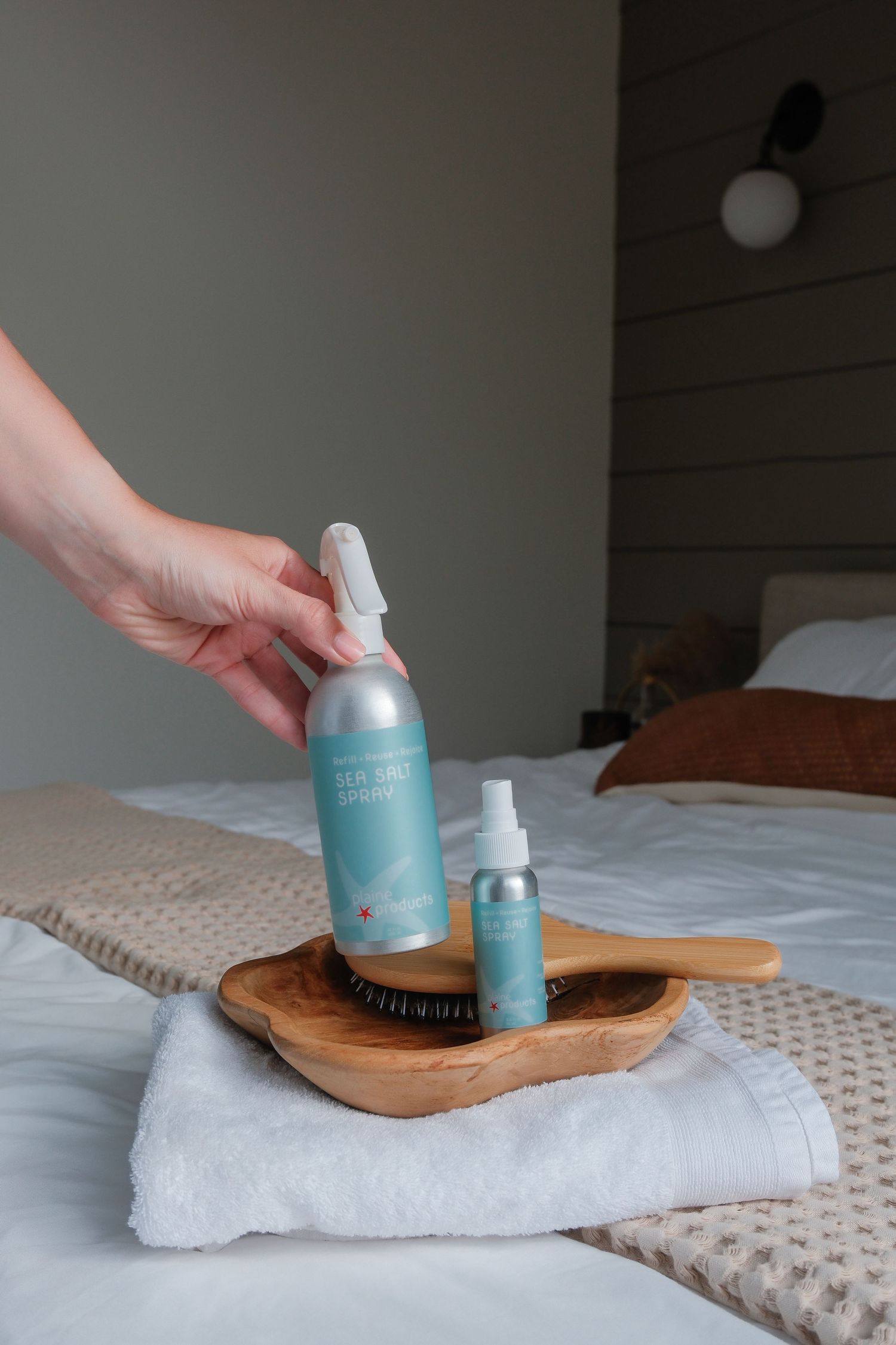 Hand holding a light blue sea salt spray bottle on a wooden tray with towel and travel size bottle.