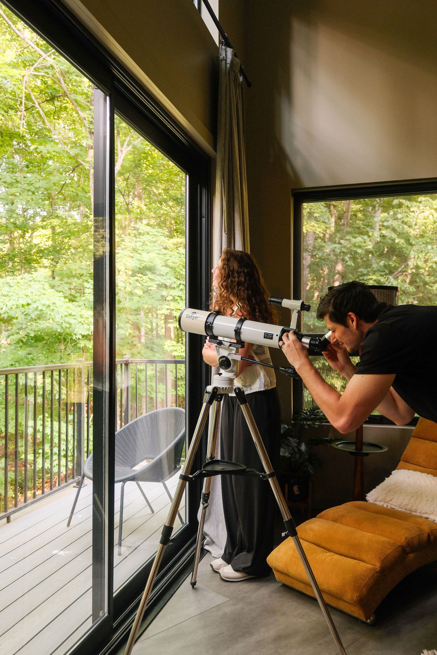 Two people using a white telescope on a tripod indoors near large windows overlooking a lush green forest.