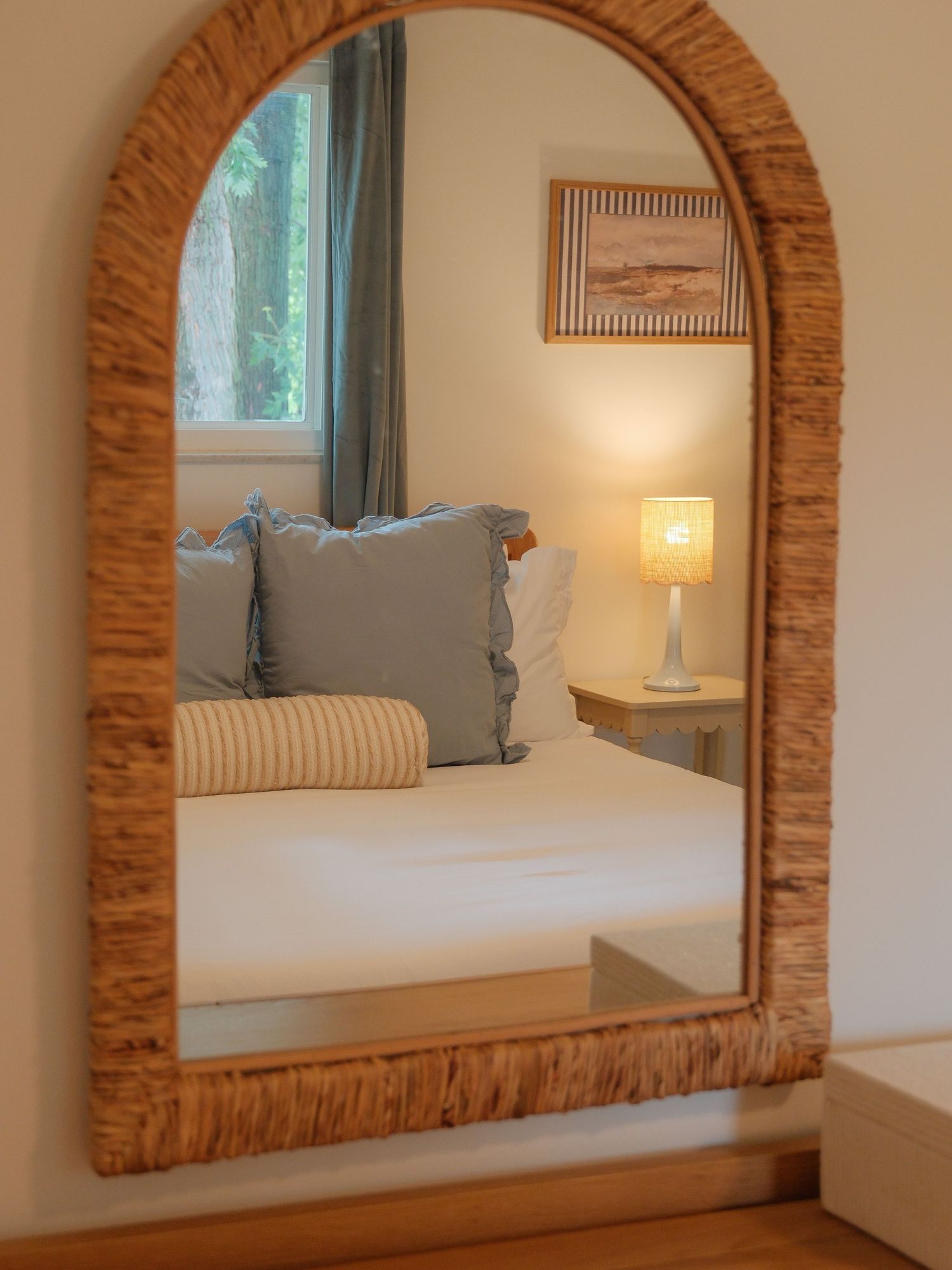 Arched rattan mirror reflecting a cozy bedroom with blue pillows, striped bedding, and a glowing bedside lamp.