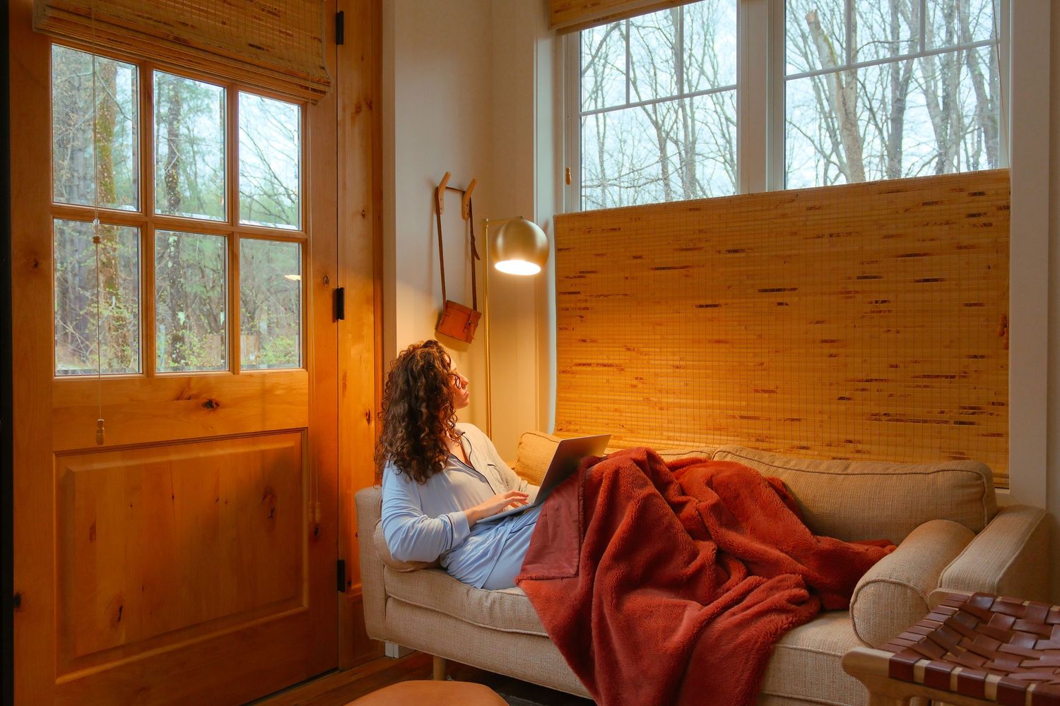Woman relaxing on a couch with a red blanket and laptop in a cozy cabin with large windows.