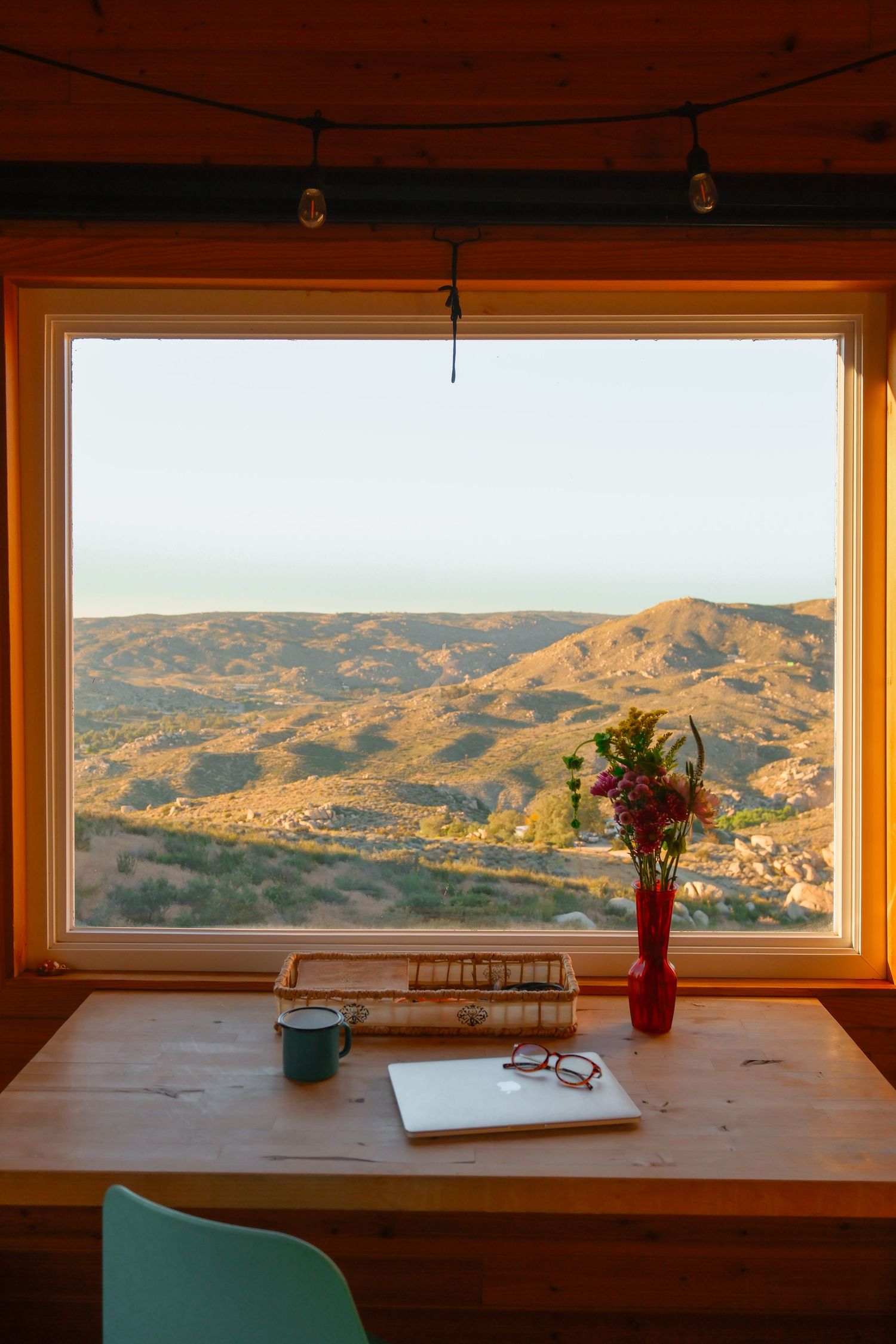 Cozy wooden desk with laptop, books, and flowers overlooking a stunning mountain valley through a large window at sunset.