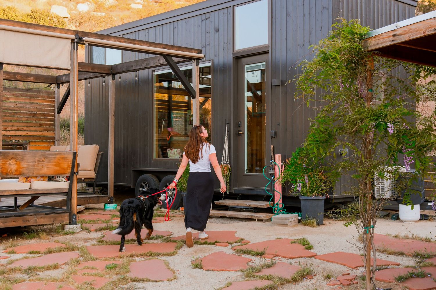 Woman walking black dog outside modern dark-clad tiny home with stepping stones and desert landscaping.