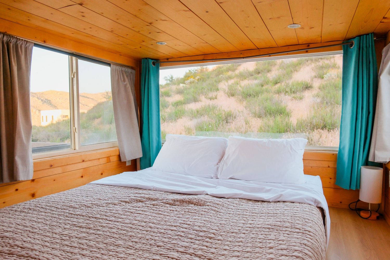 Cozy cabin bedroom with wood-paneled walls, large window, teal curtains, and a queen bed with desert hill views.