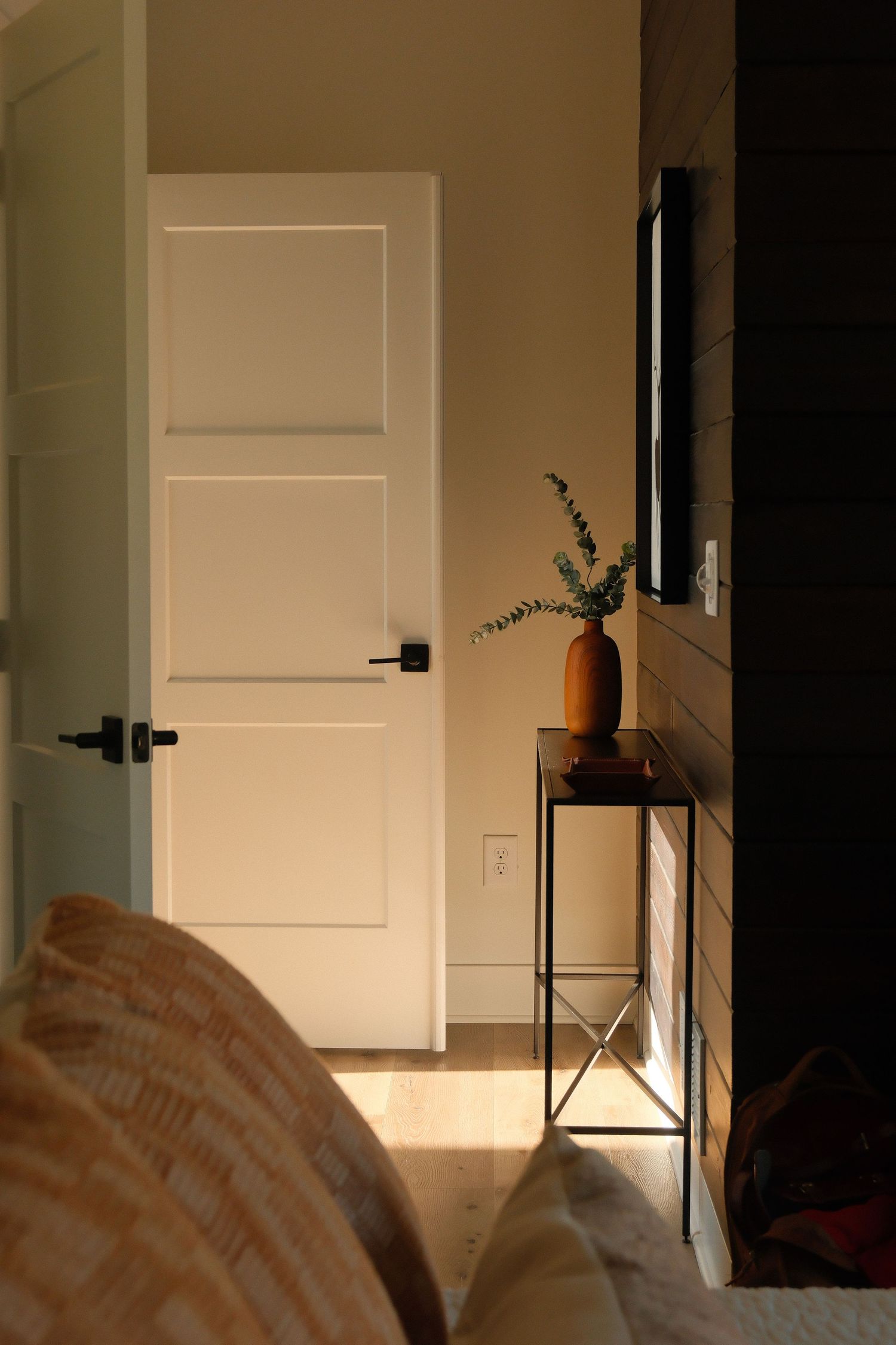 Cozy bedroom corner with white paneled door, metal side table, and terracotta vase with eucalyptus in warm evening light.
