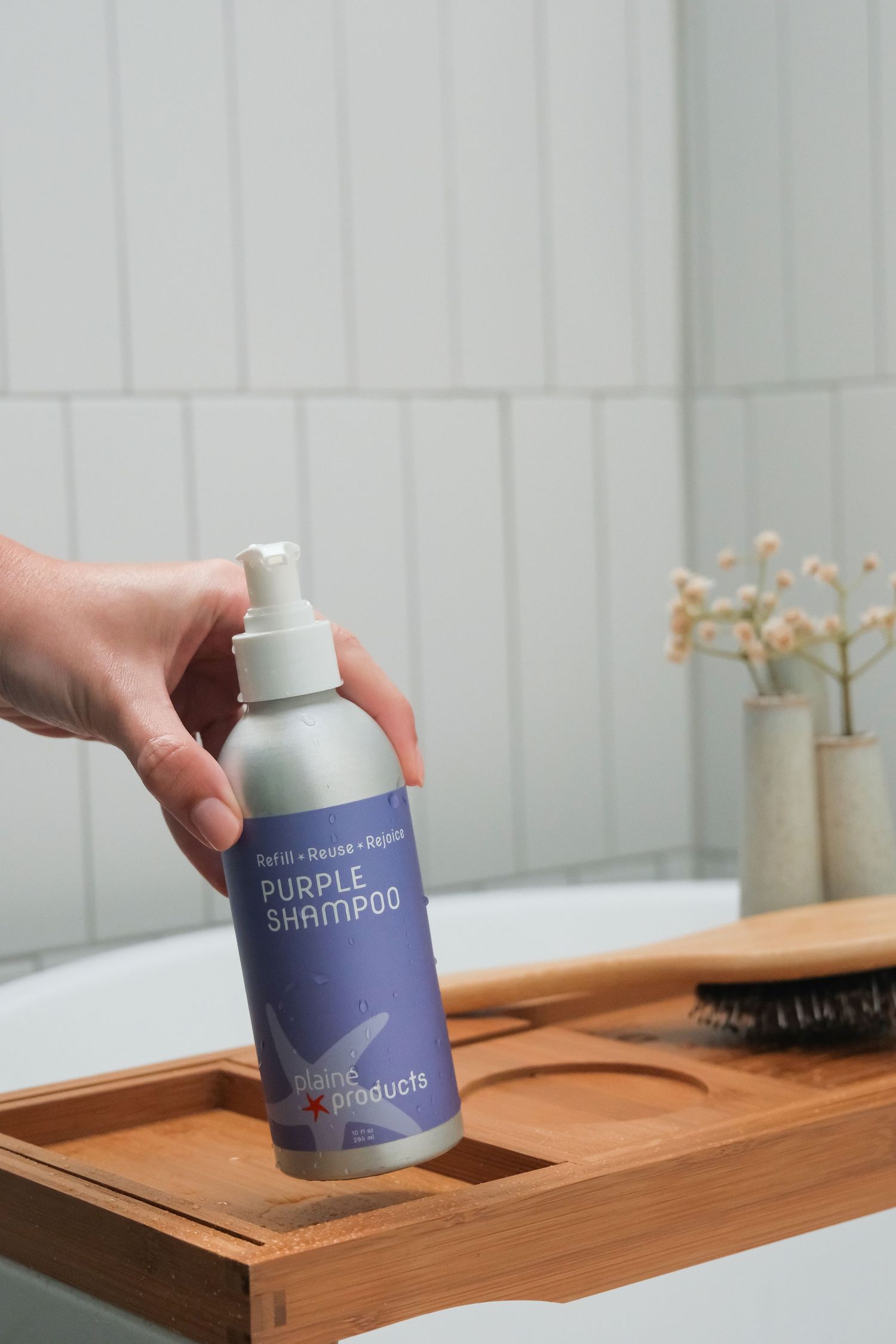 Hand holding a aluminum bottle of Blank Products Purple Shampoo on a wooden bath tray in a modern bathroom.