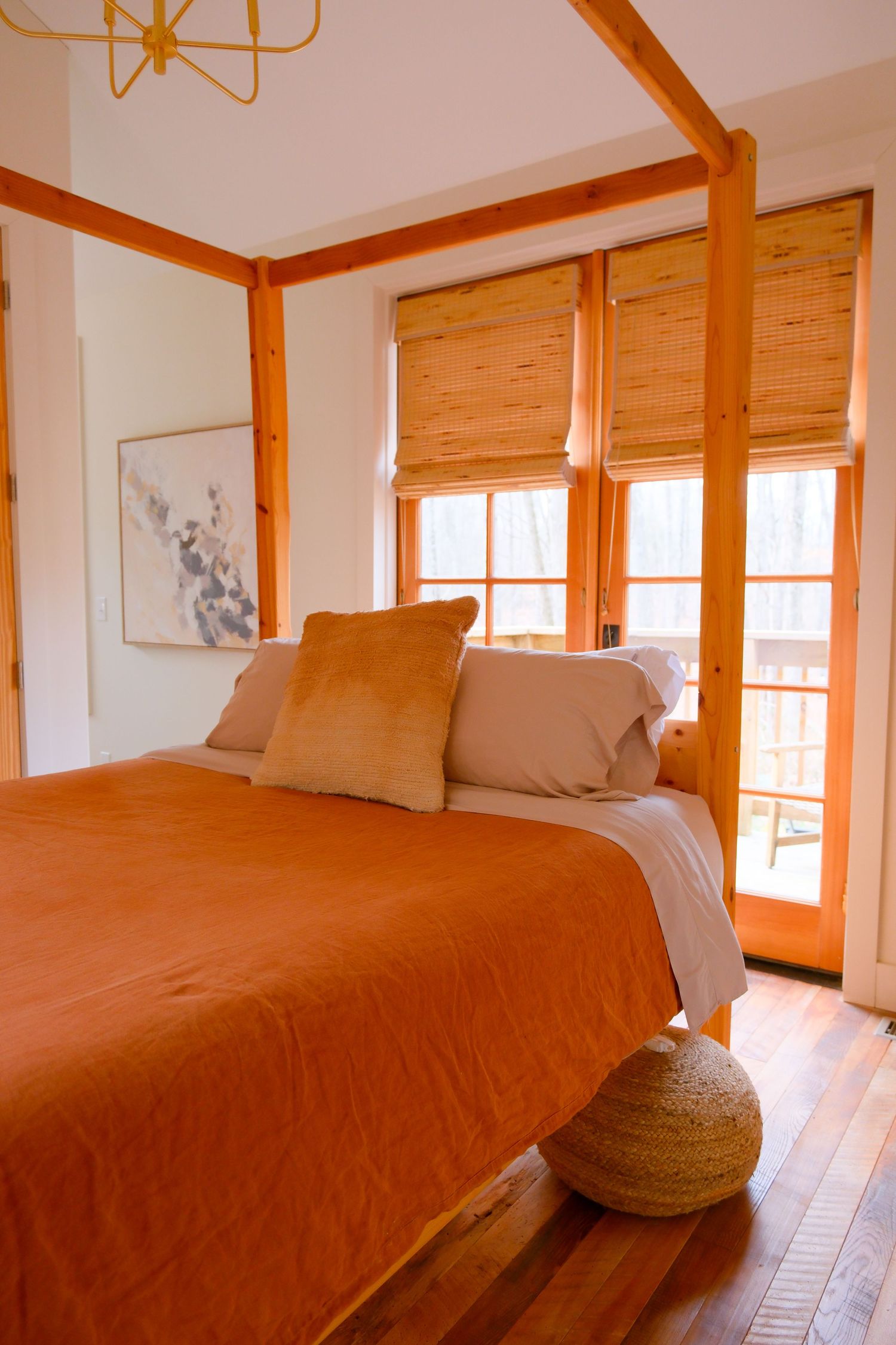 Cozy bedroom with orange bedding, wooden four-poster frame, bamboo shades, and hardwood floors bathed in warm natural light.