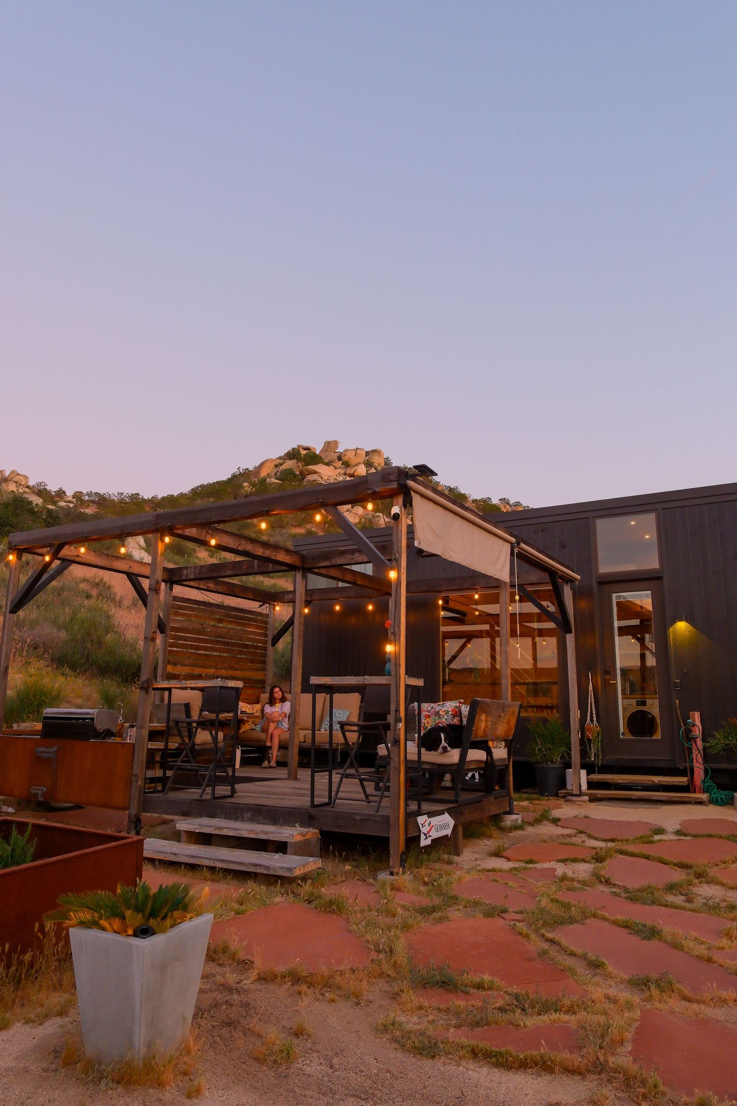 Rustic outdoor pergola with string lights at dusk, nestled against rocky hills near a modern dark cabin.