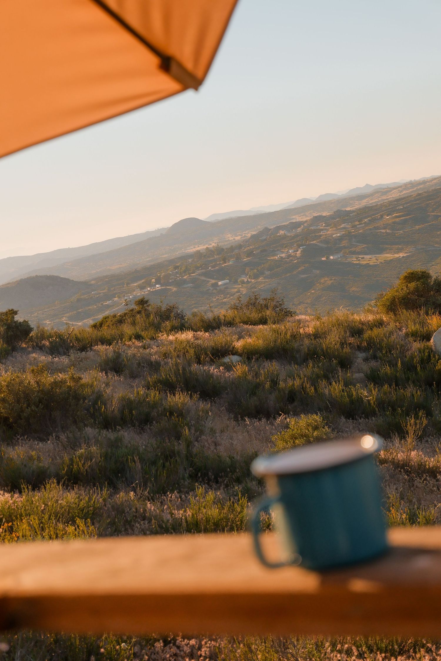 A blue enamel camping mug sits on a wooden ledge overlooking a scenic mountain landscape at golden hour.