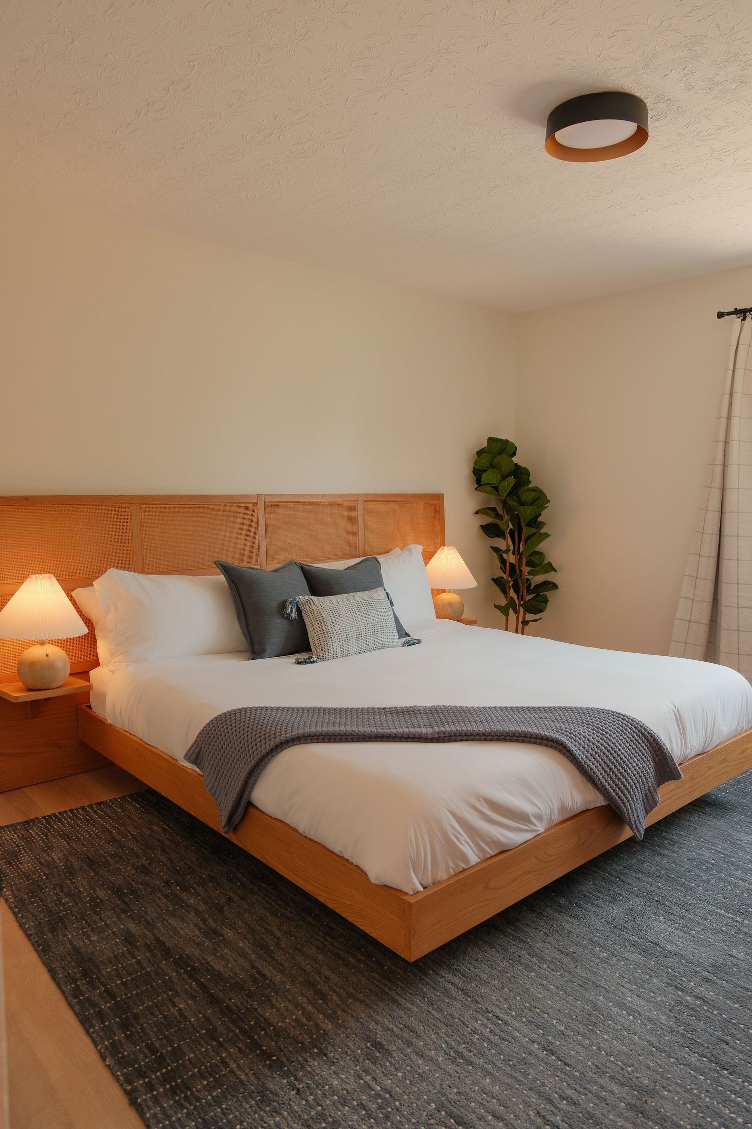 Modern bedroom with warm wood bed frame, white bedding, gray throw, matching nightstands, and a fiddle leaf fig plant.
