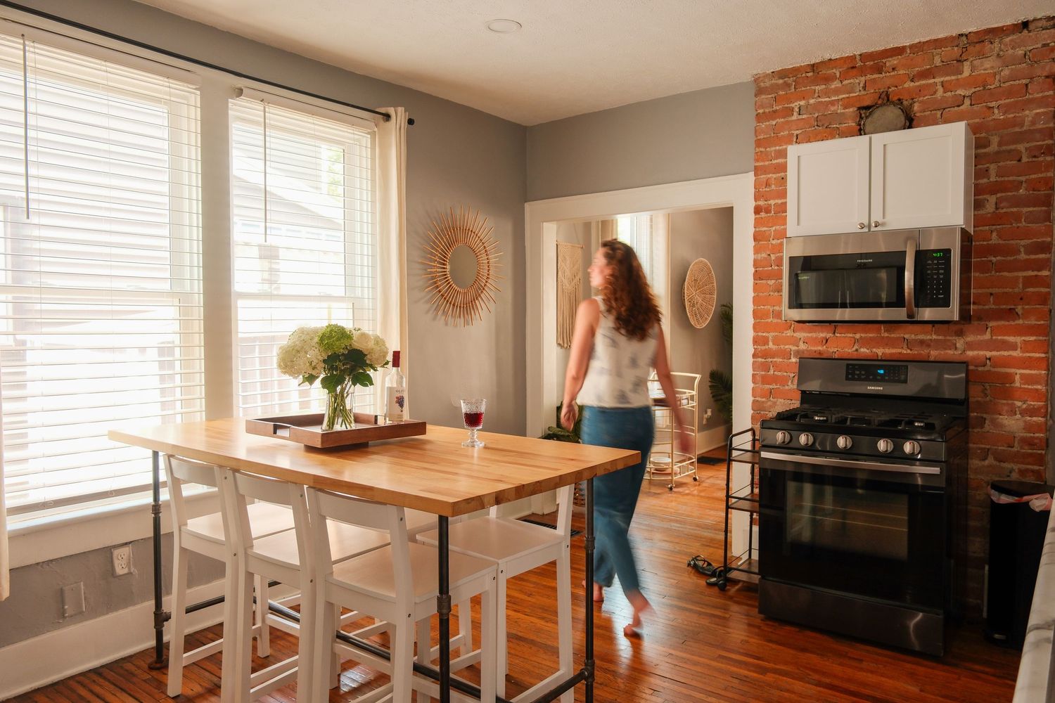 Woman walking through a modern kitchen with brick accent wall, gas stove, and dining table with wine glass.