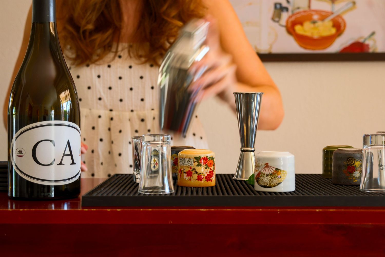 Bartender shaking cocktails beside a CA wine bottle and decorative cups on a bar counter.