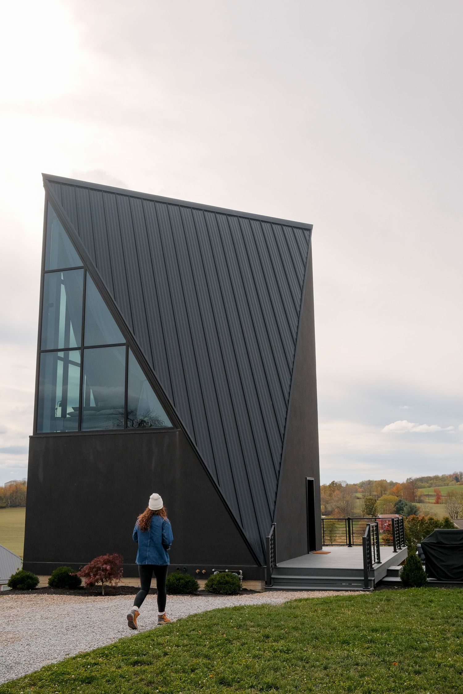 Woman in blue jacket walks toward modern angular black building with geometric facade on a cloudy day.