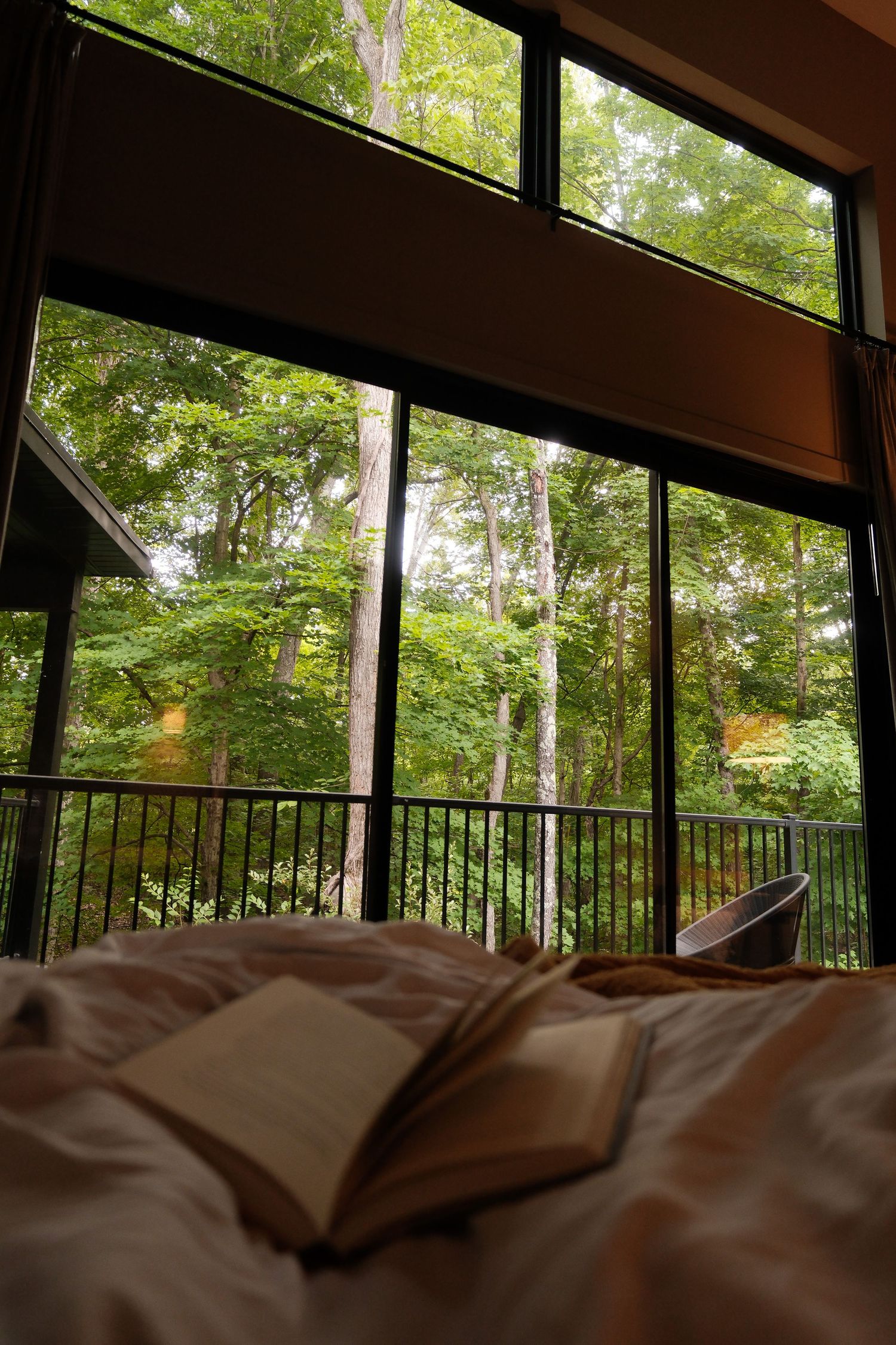 Open book on a bed with a serene forest view through large floor-to-ceiling windows on a lush green day.
