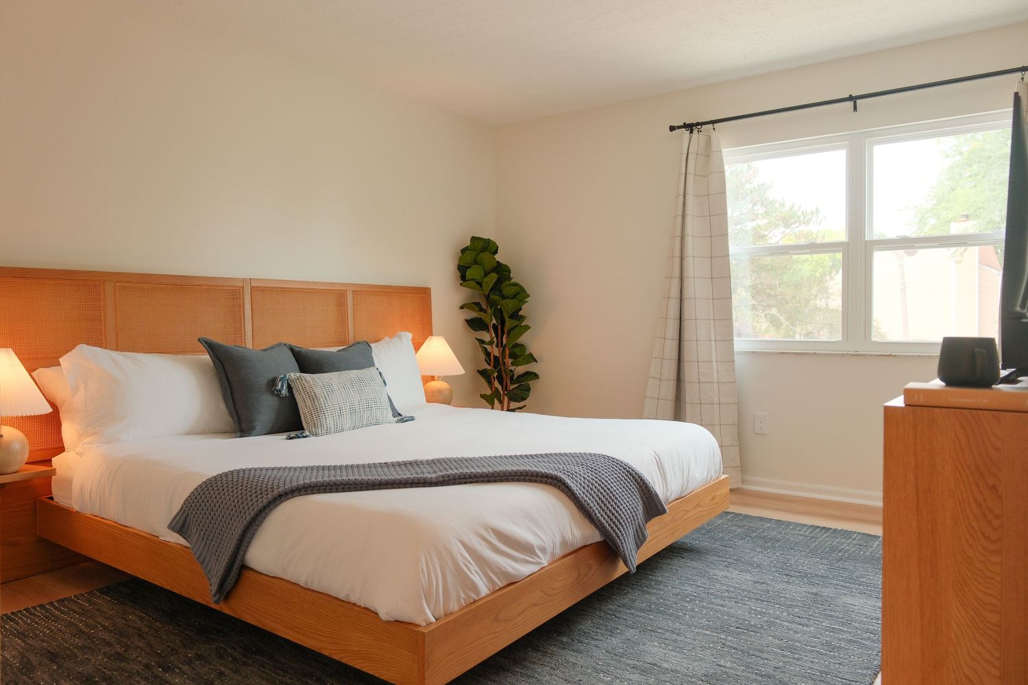 Modern bedroom with warm wood bed frame, gray accent pillows, plush bedding, blue rug, and natural light from window.