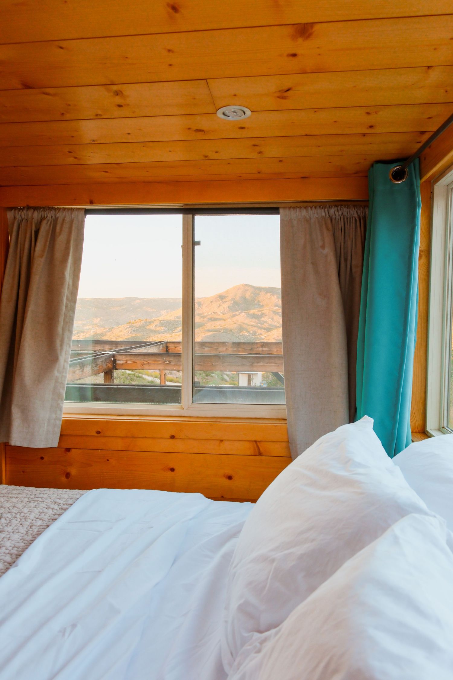 Cozy wooden cabin bedroom with white pillows, teal curtains, and a scenic mountain view through the window.