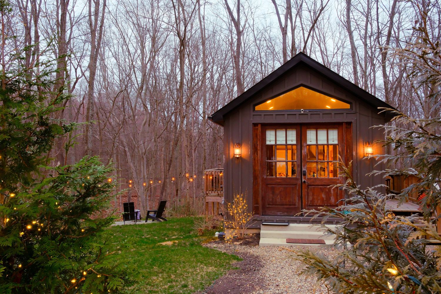 Cozy rustic cabin nestled in bare winter woods, glowing with warm lights and string lights at dusk.
