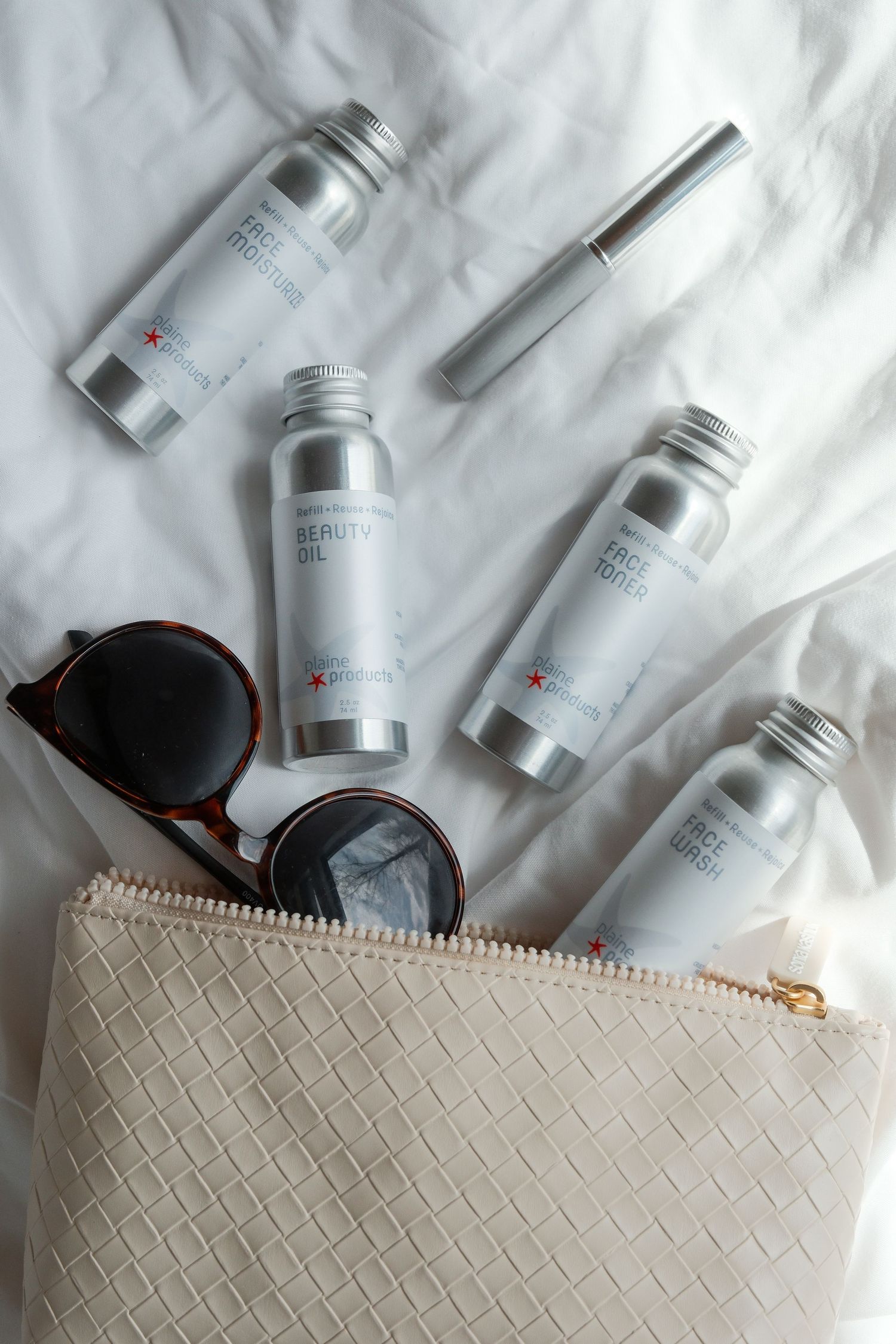 Flat lay of silver travel-size skincare bottles spilling from white woven cosmetic bag with sunglasses on white bedding.