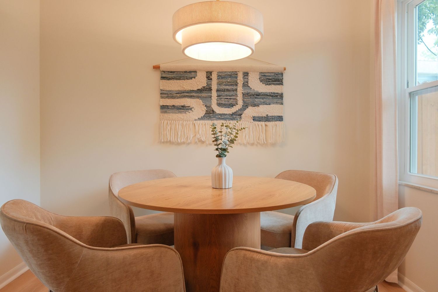 Cozy dining area with round wood table, beige chairs, drum pendant light, and woven wall art.
