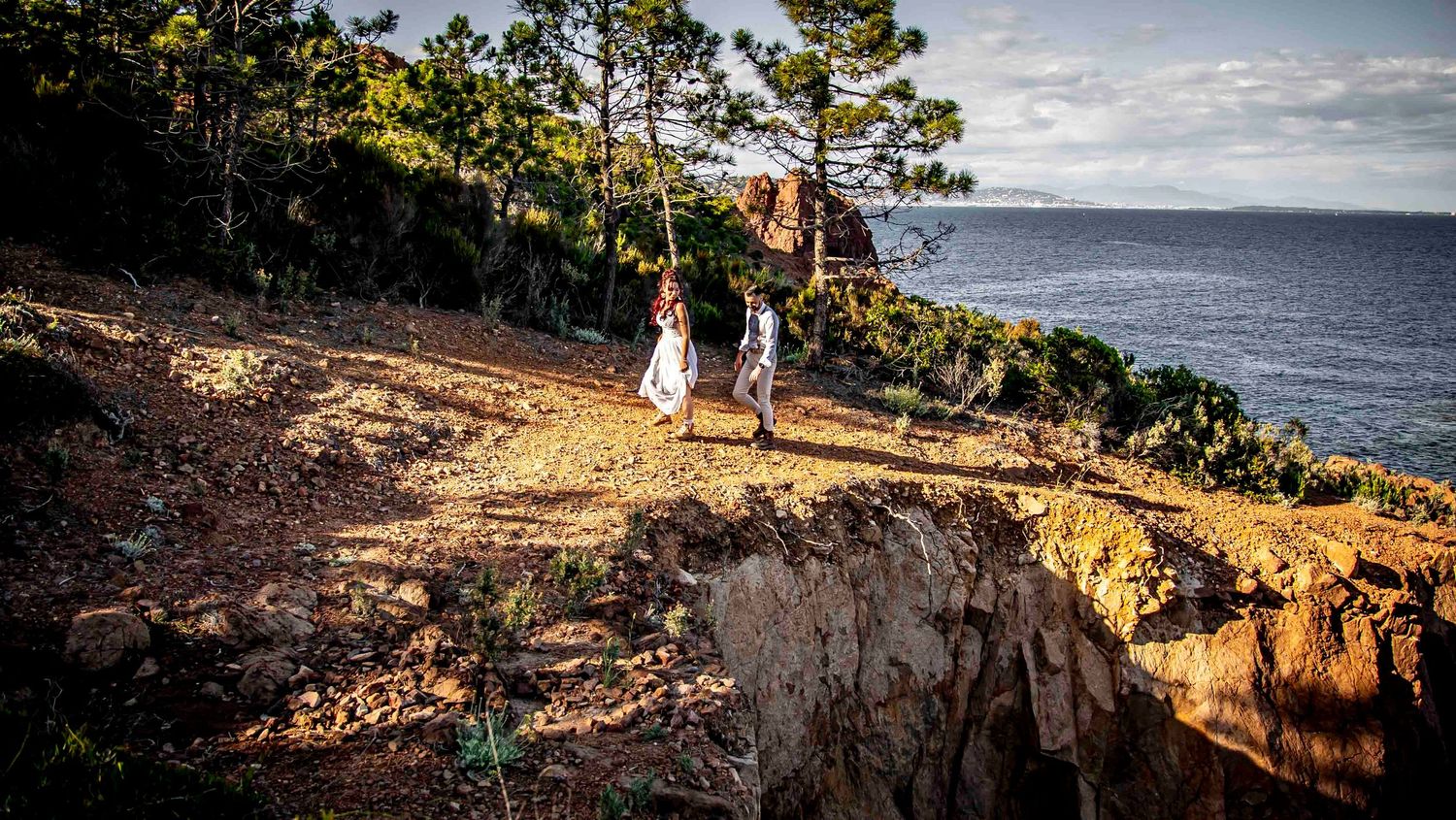 Hikers walk along a scenic coastal cliff trail lined with pine trees overlooking the ocean on a sunny afternoon.