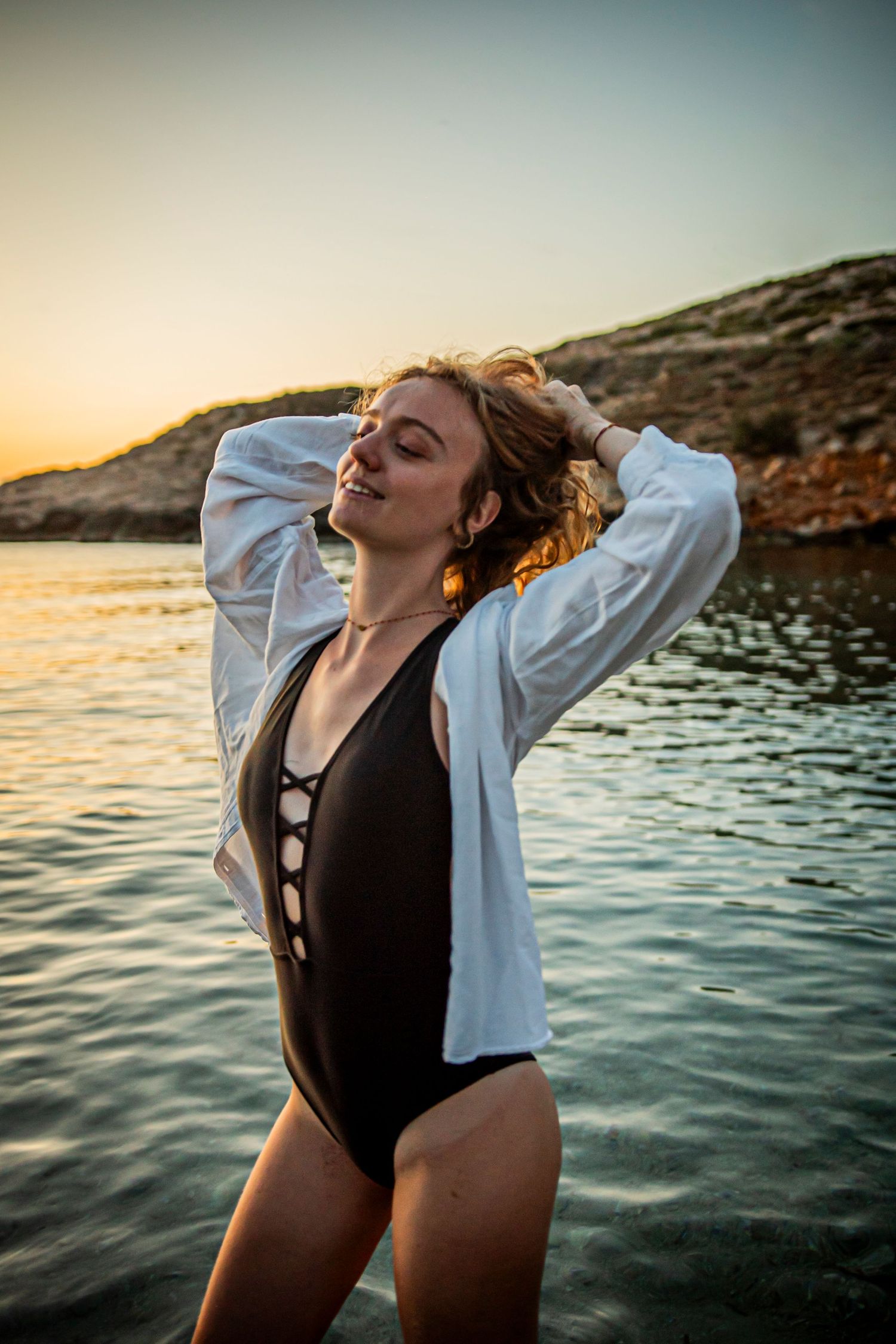 Woman in swimsuit enjoying sunset by rocky coastline with golden light.