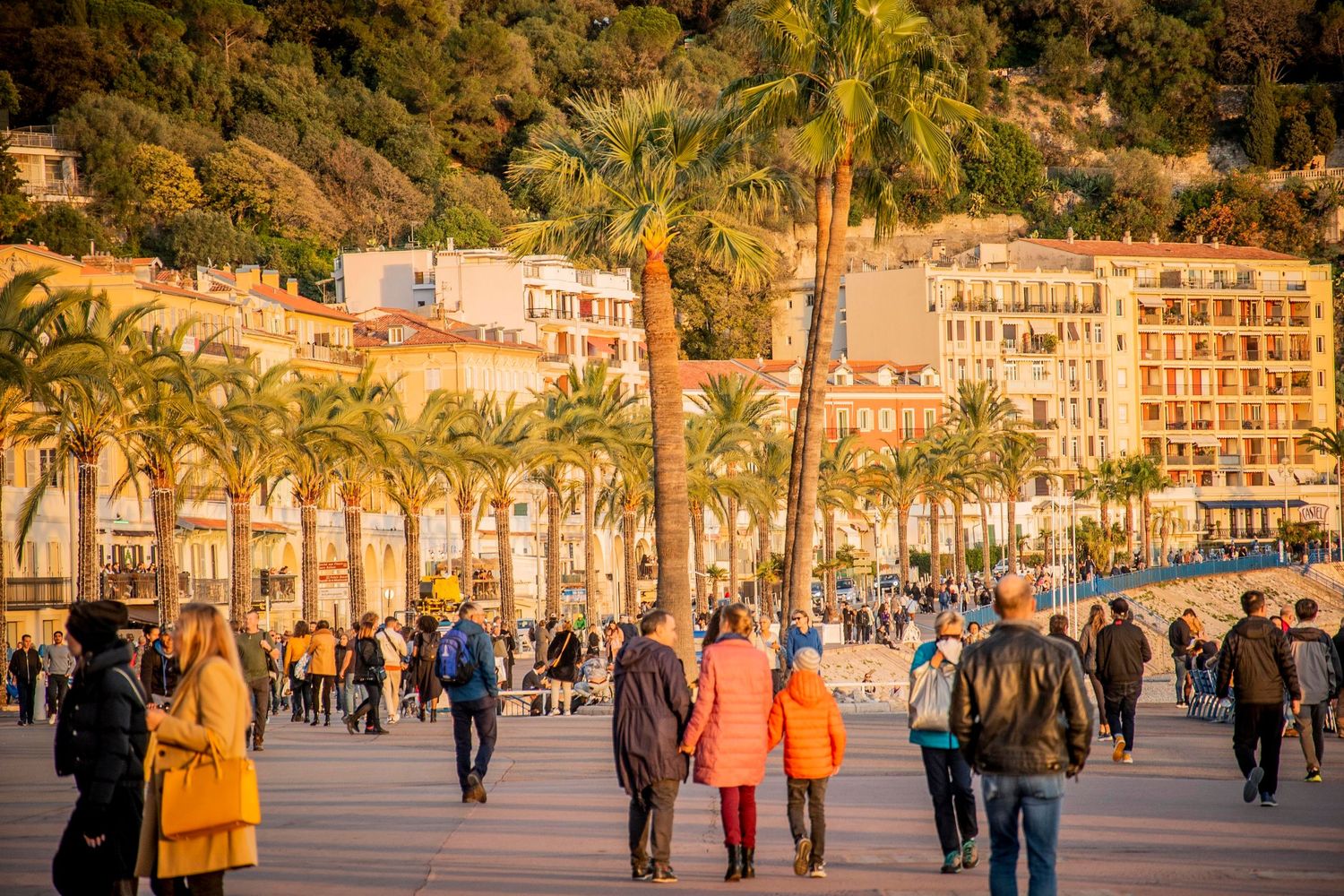 Tourists stroll along a palm-lined waterfront promenade in a Mediterranean coastal city during golden hour.