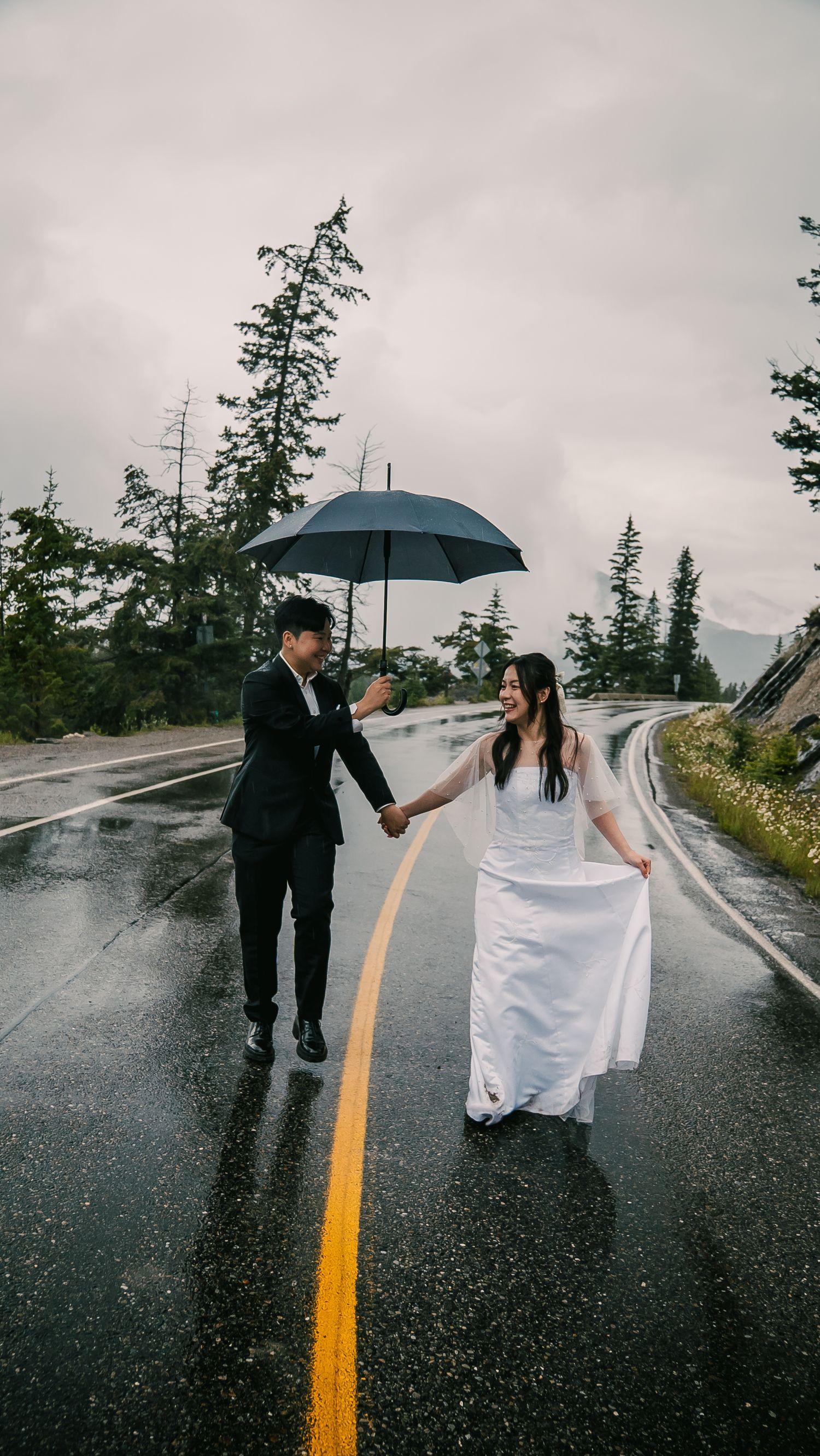 A couple in wedding attire hold hands on a wet mountain road under an umbrella on a rainy, misty day.