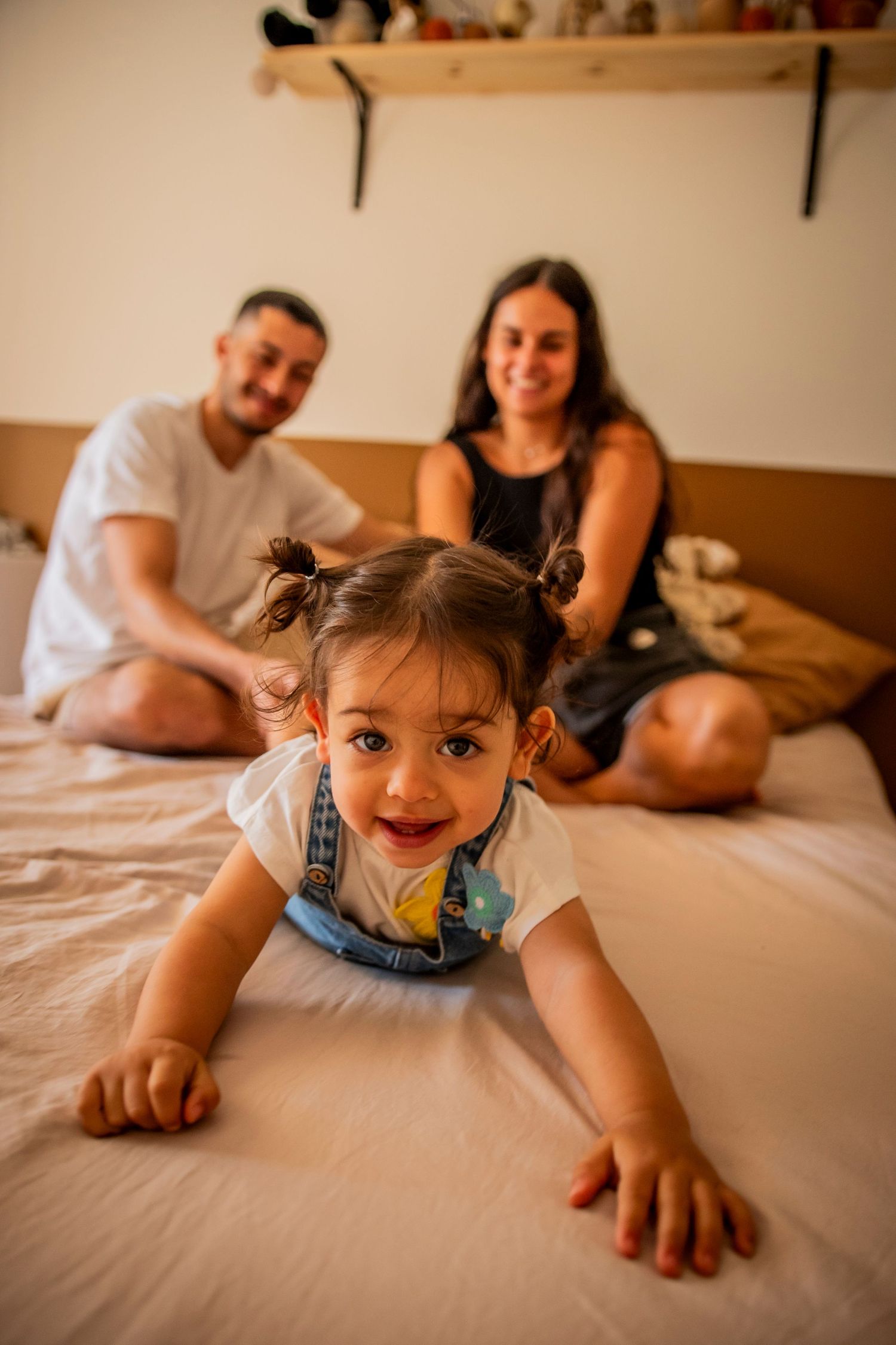 Family relaxing together on white bed with small child crawling playfully.