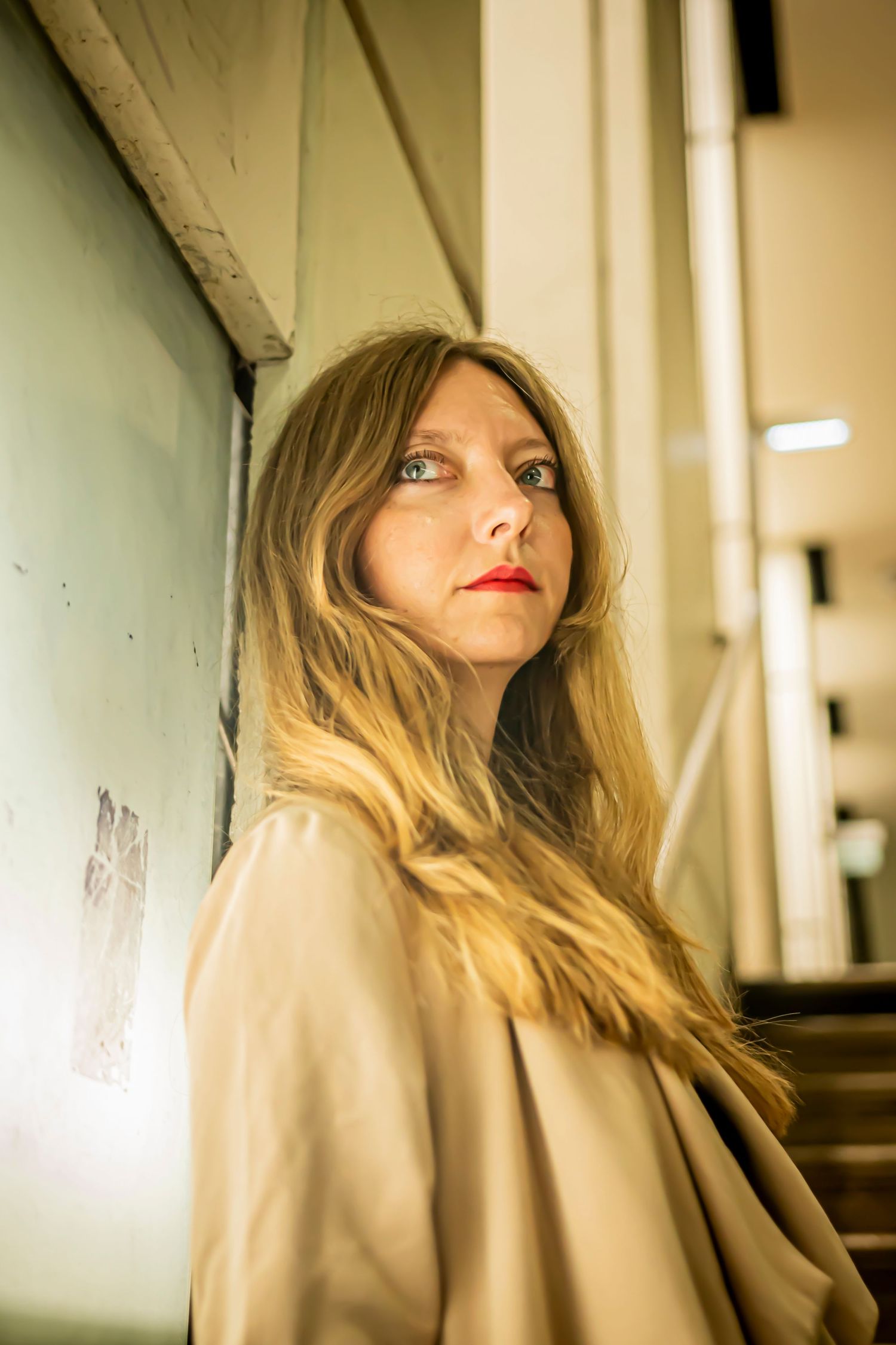 Stylish woman with long blonde hair and red lipstick posing against wall in beige coat.