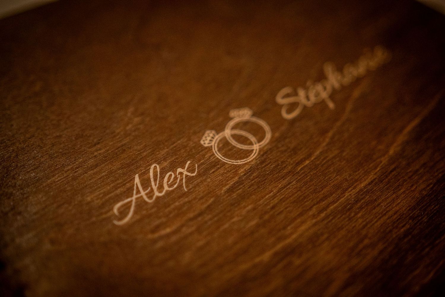 Wedding rings and names engraved in elegant script on a dark wooden surface.