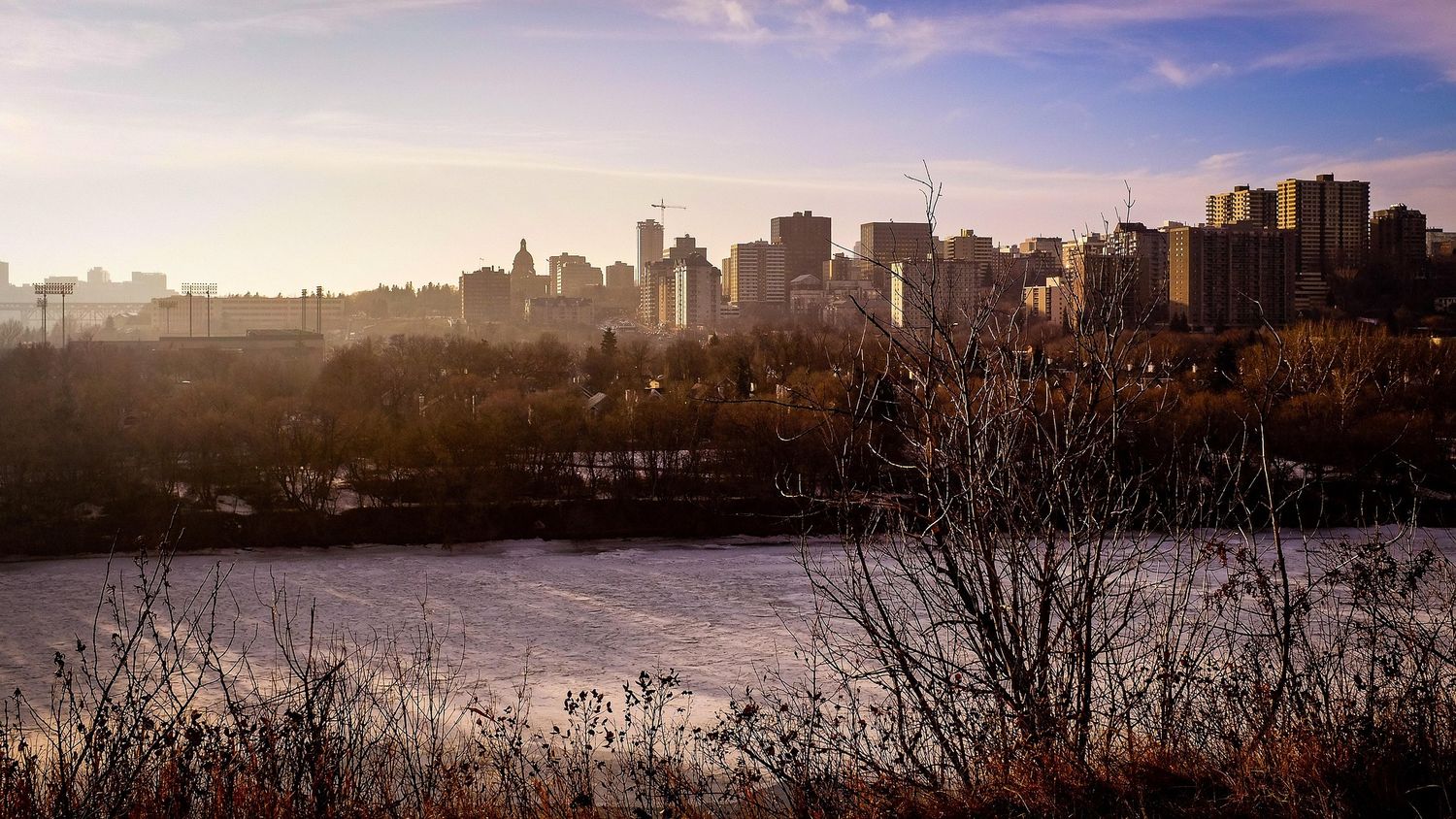 Winter sunset view of Edmonton skyline with frozen North Saskatchewan River and bare trees in foreground.