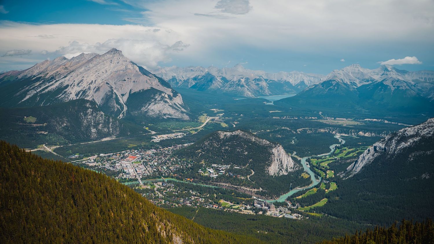 Panoramic mountain landscape view showing dramatic peaks and valleys under a bright blue sky with scattered clouds.