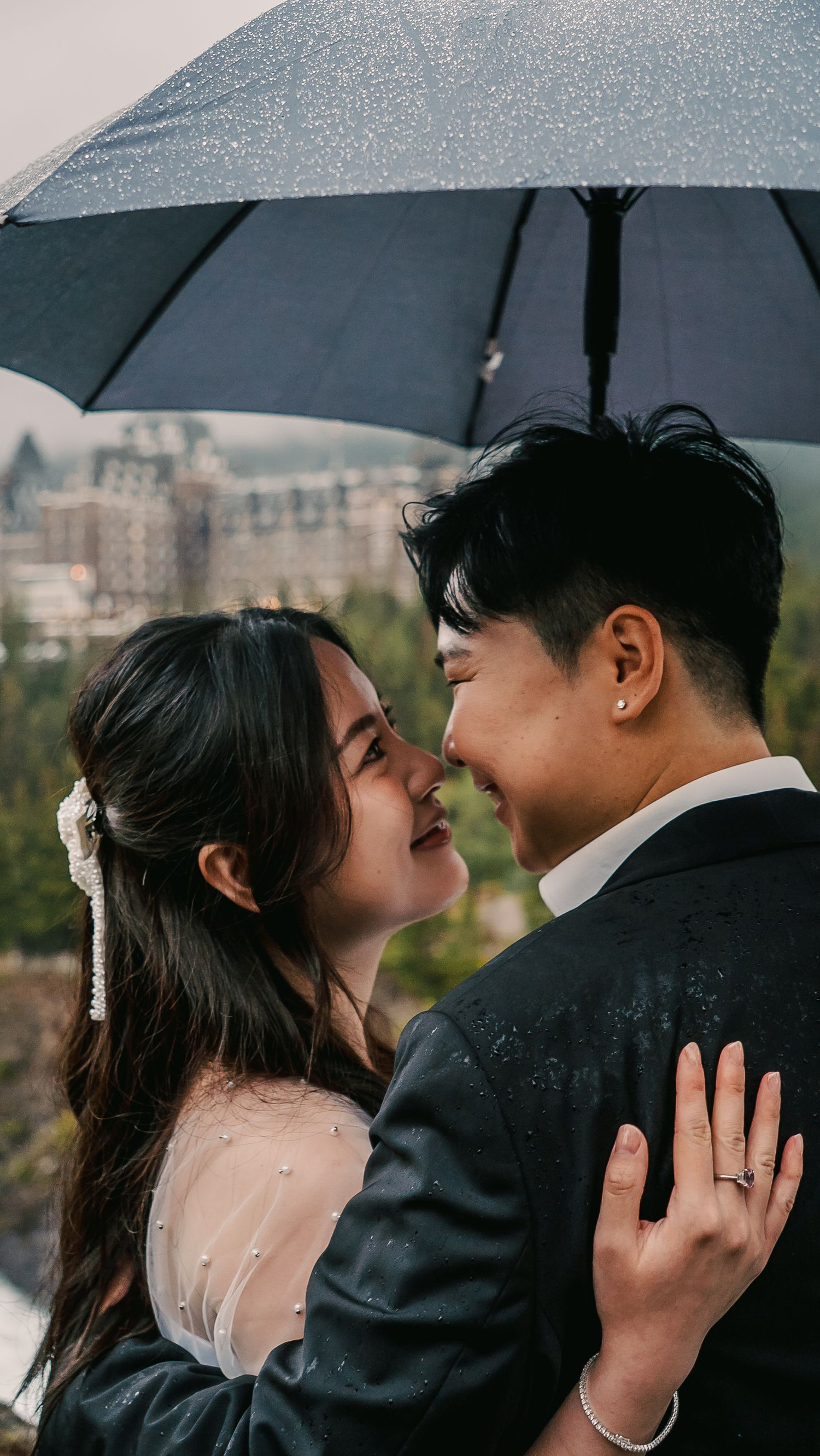 A romantic couple in formal attire share intimate moments under a black umbrella on a rainy day.