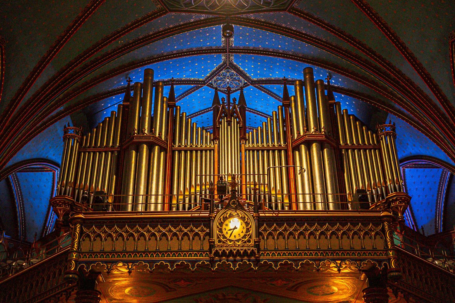 Illuminated golden pipe organ with ornate Gothic architectural details glows against deep blue stained glass in a cathedral.