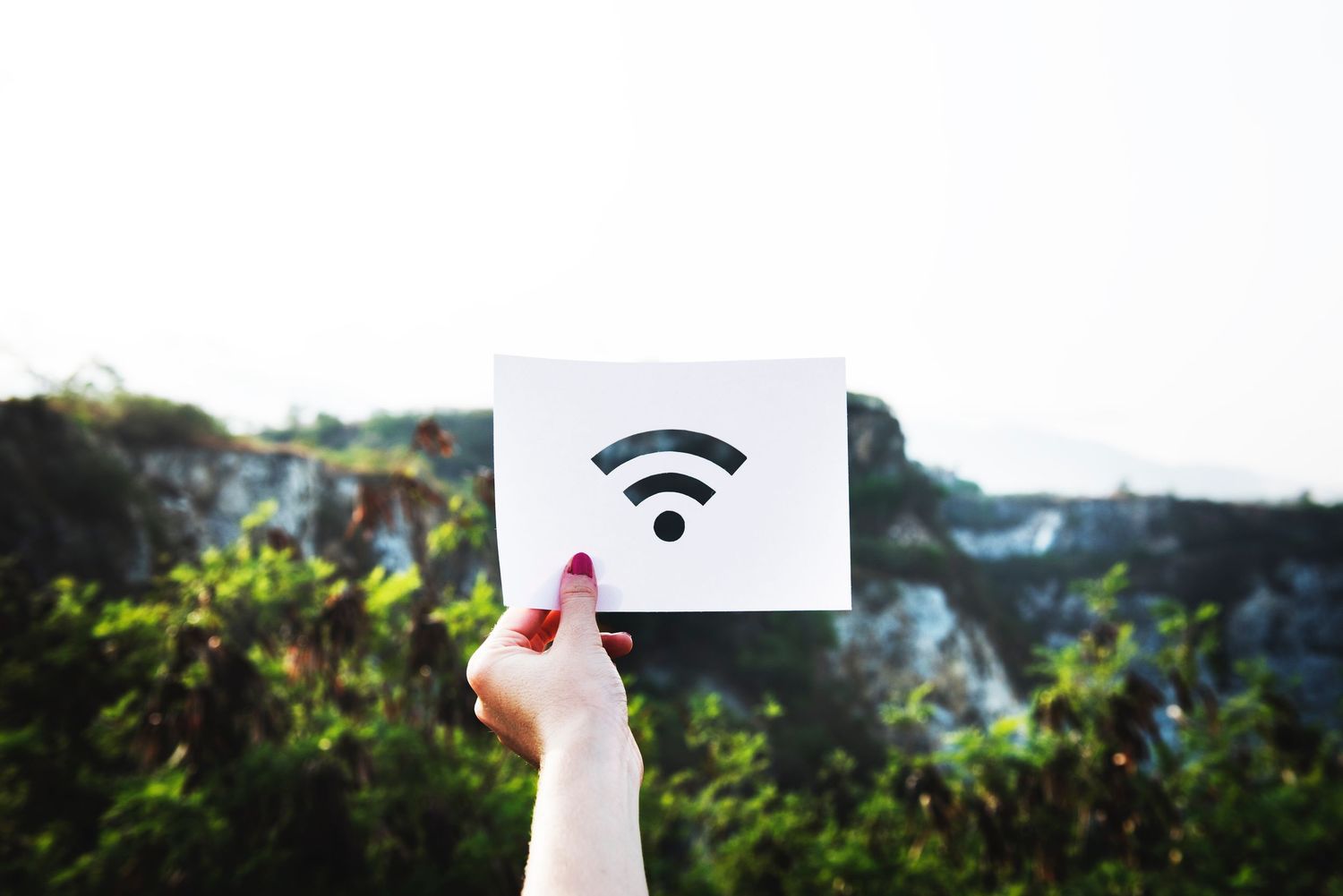 Hand holding up a white paper with wifi symbol against green outdoor background with trees and sky.