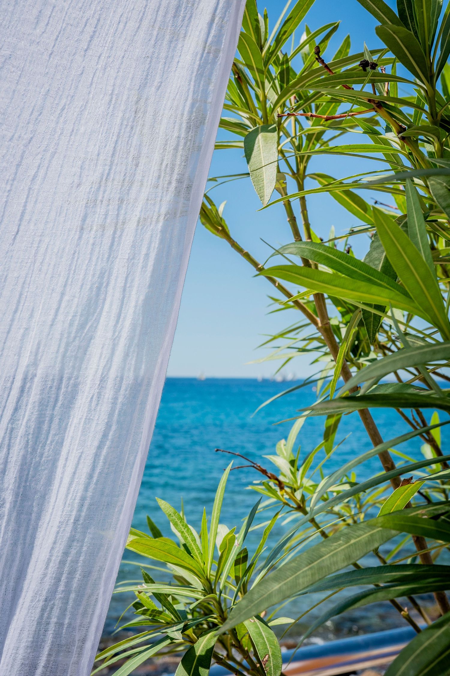 White curtain and tropical plants frame view of bright blue Mediterranean sea.
