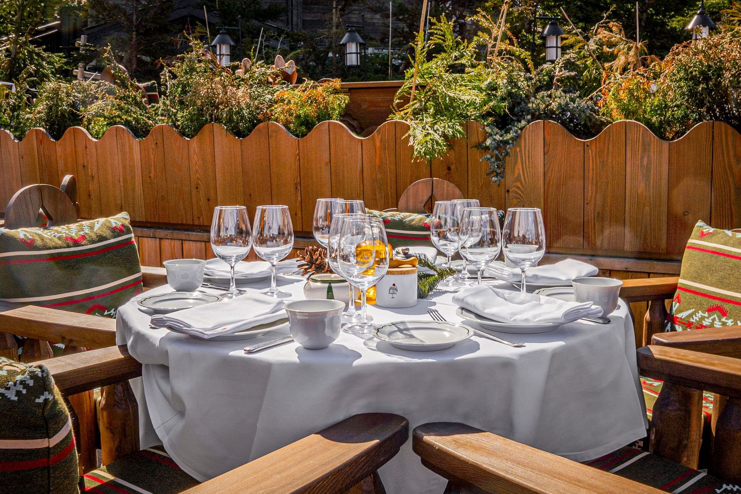 Elegant outdoor dining table set with white tablecloth, wine glasses and place settings on wooden patio with garden backdrop.
