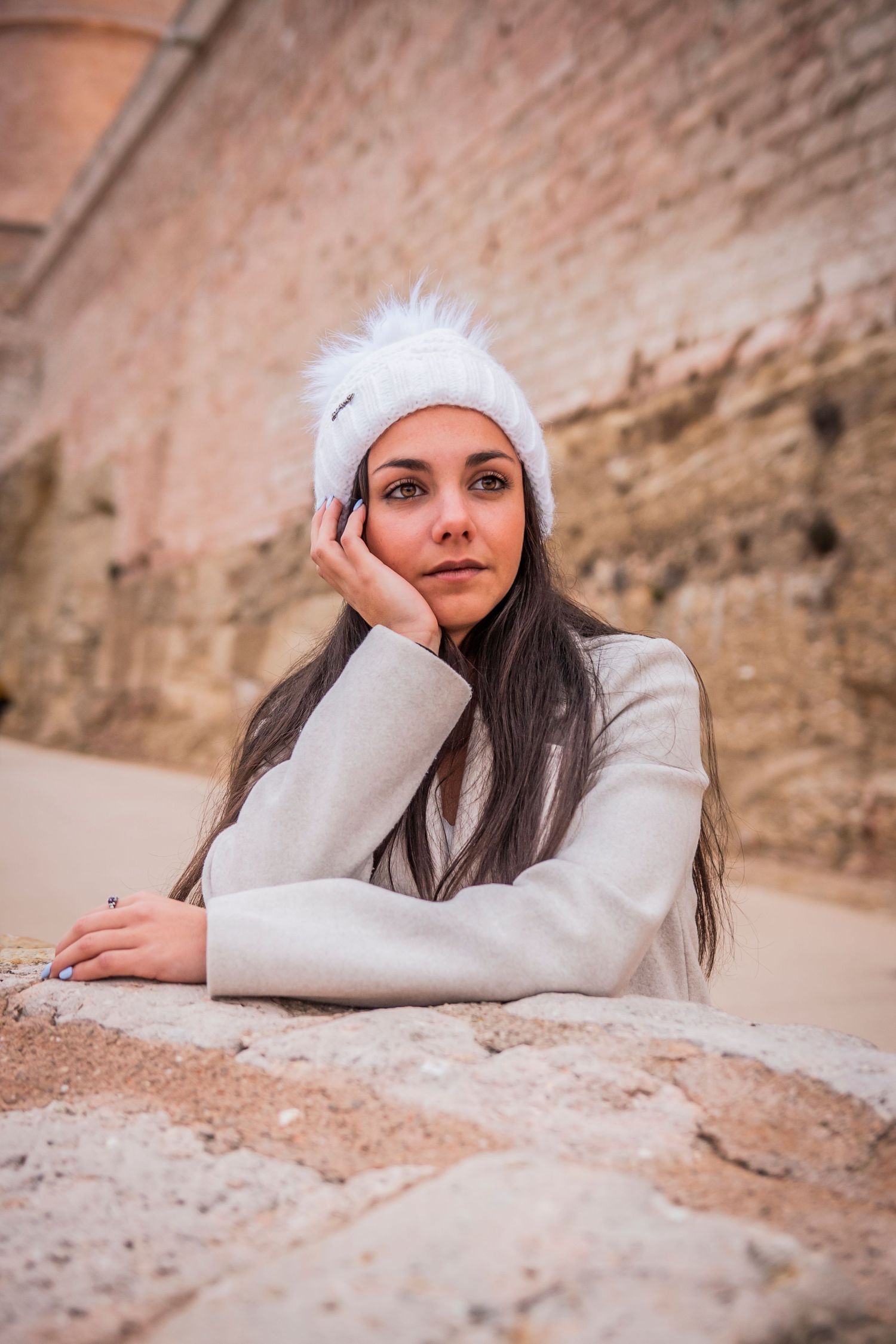 A person in a white winter hat poses thoughtfully against a brick wall.