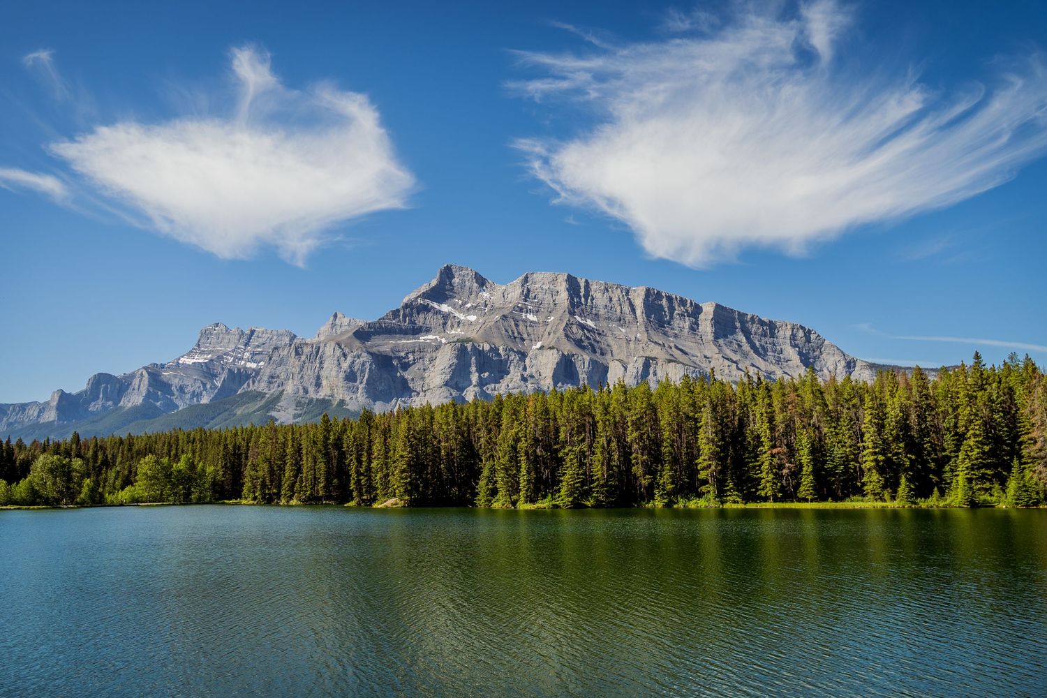 Majestic Mount Rundle towers over Two Jack Lake surrounded by pine forest in Banff National Park on a sunny day.